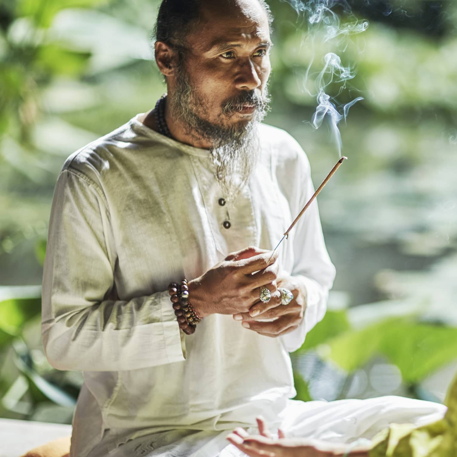 Set against a backdrop of greenery, Djik Dewa holds a stick of burning incense before a meditative cross-legged guest.