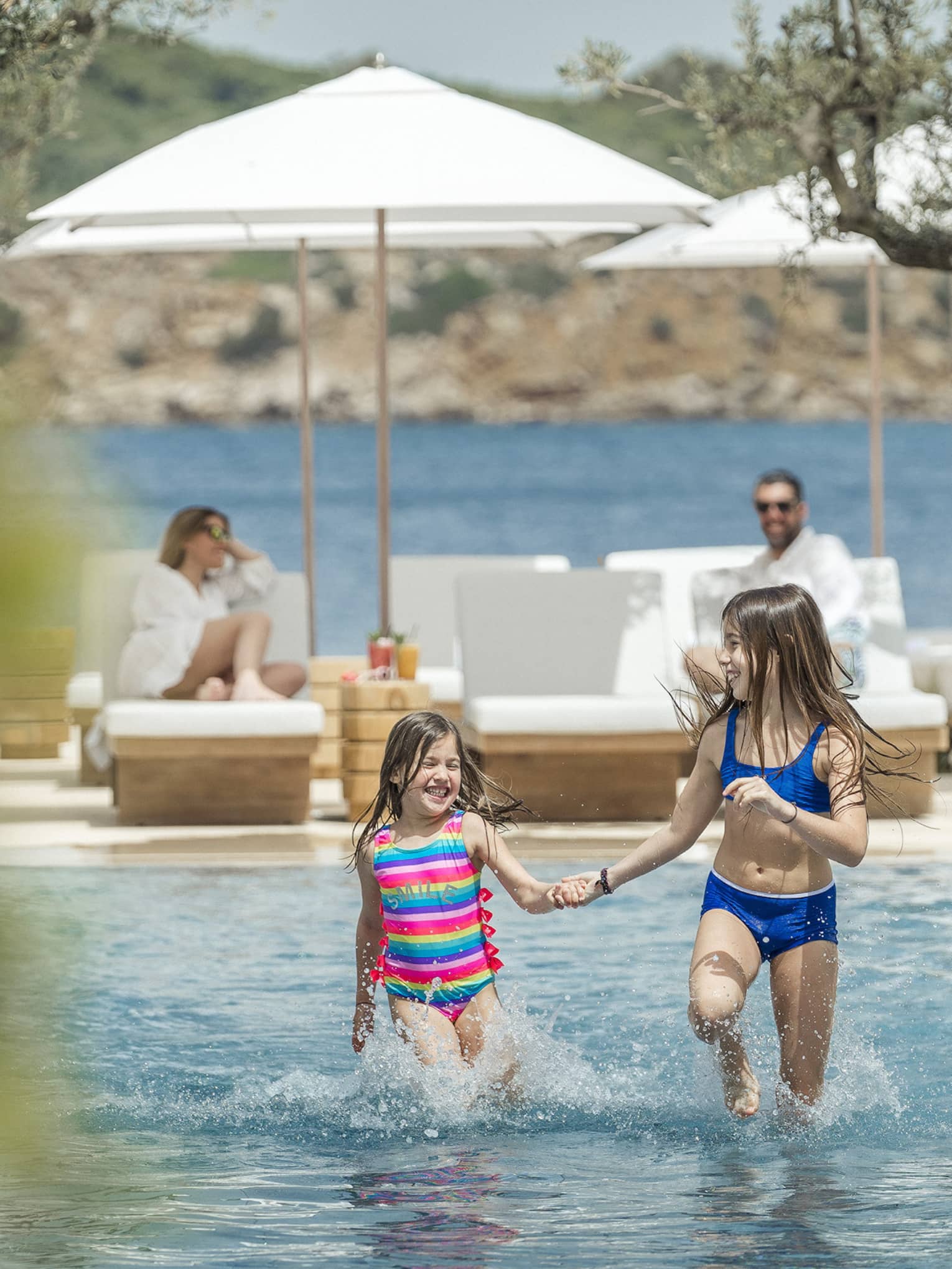 Two smiling children frolick in a shallow outdoor pool while their parents watch from sun loungers. The ocean is behind them.