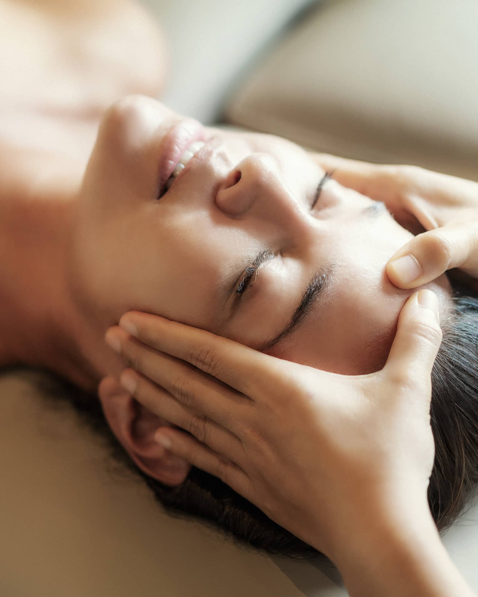 Spa facial, two hands rest on woman's forehead as she closes her eyes, lays under sheet on treatment table