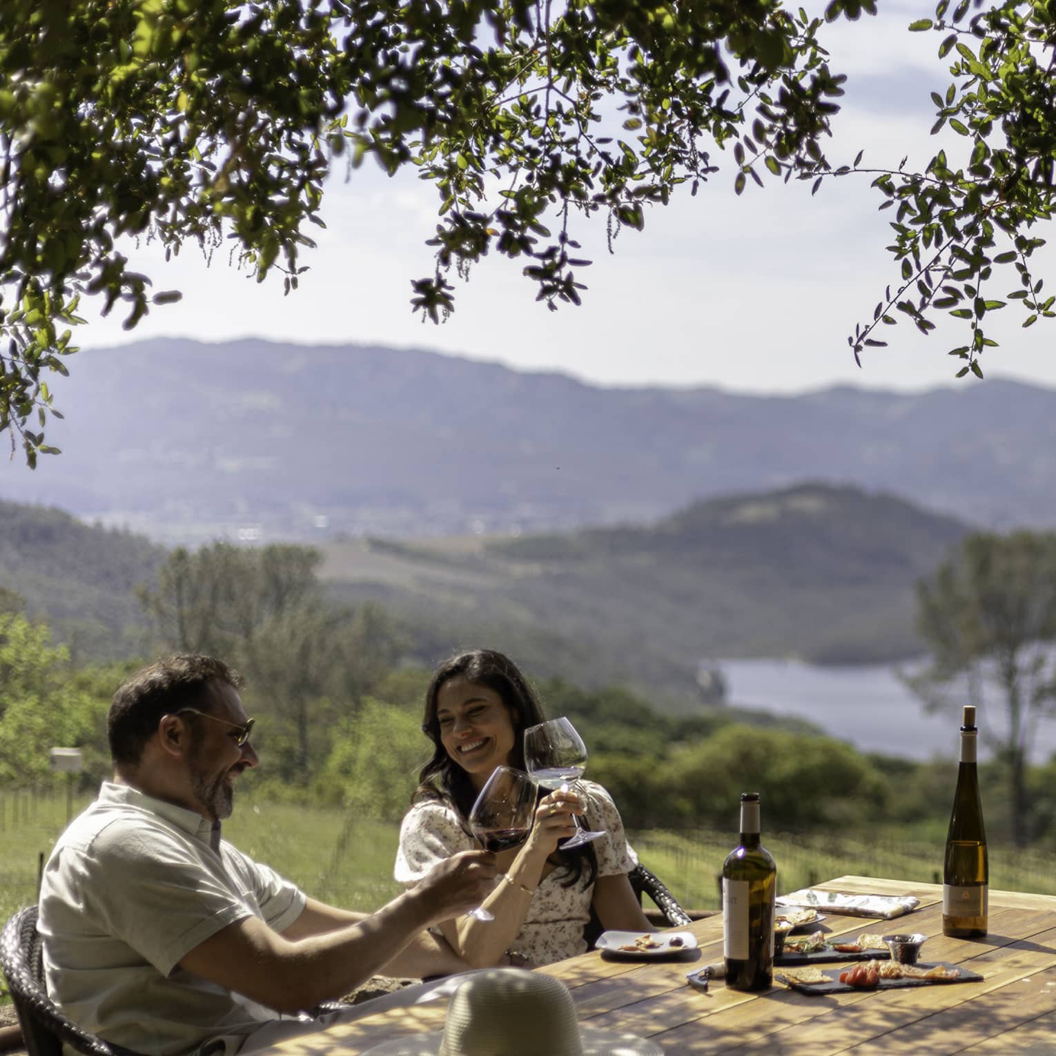 A couple smile and clink wine glasses as they sit at a rustic outdoor table overlooking a green valley and hill-lined lake.