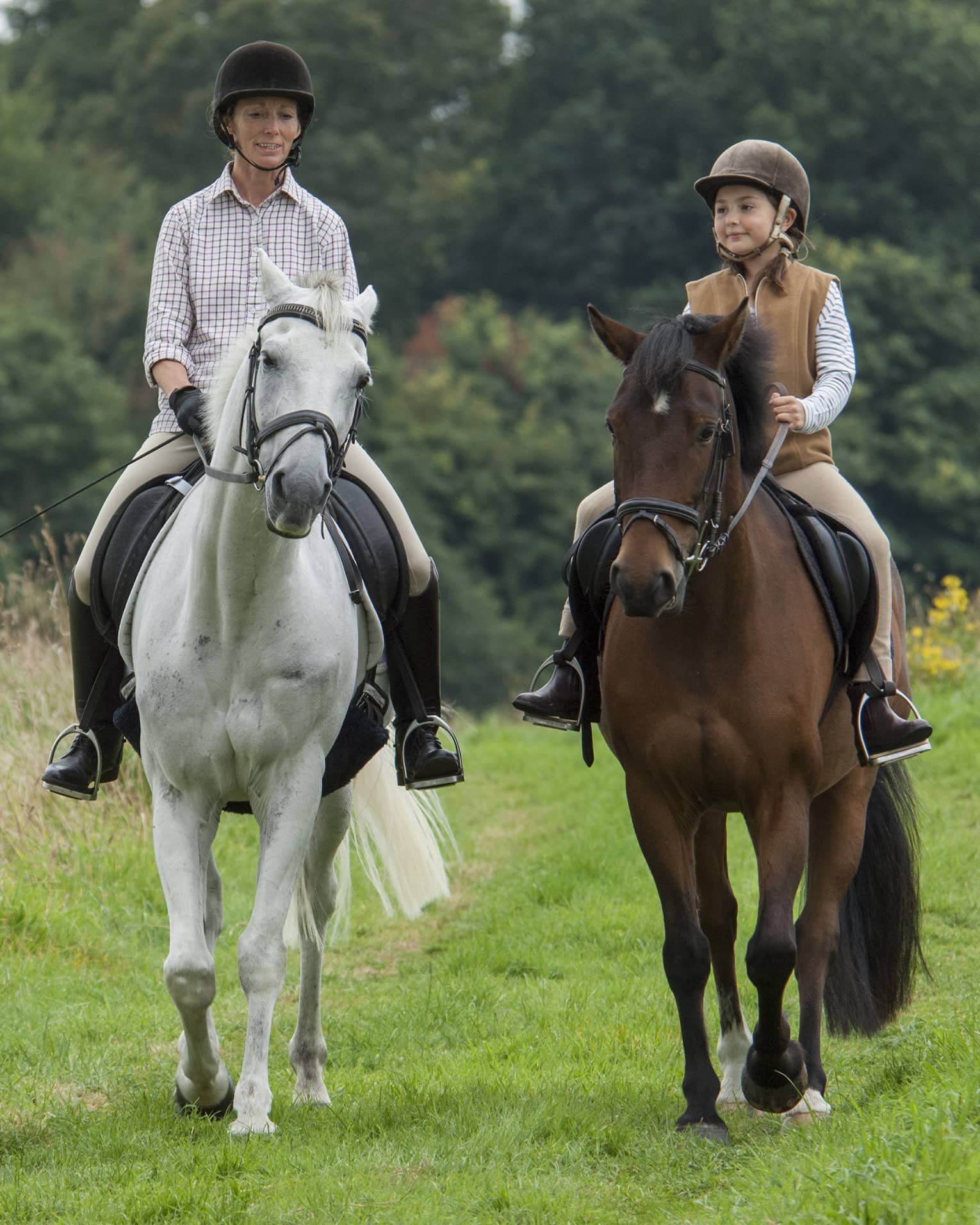 Front view of an adult and child riding horses along a trail in a verdant meadow, a forest of trees in the background.