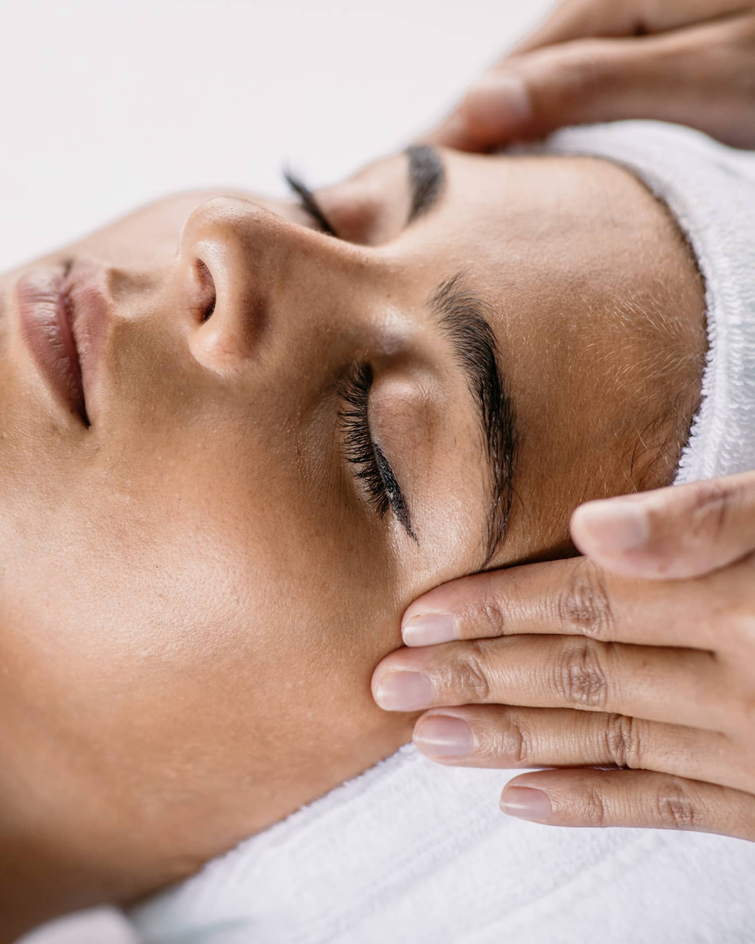 Hands massage woman's temples as she closes her eyes, spa towel around hair