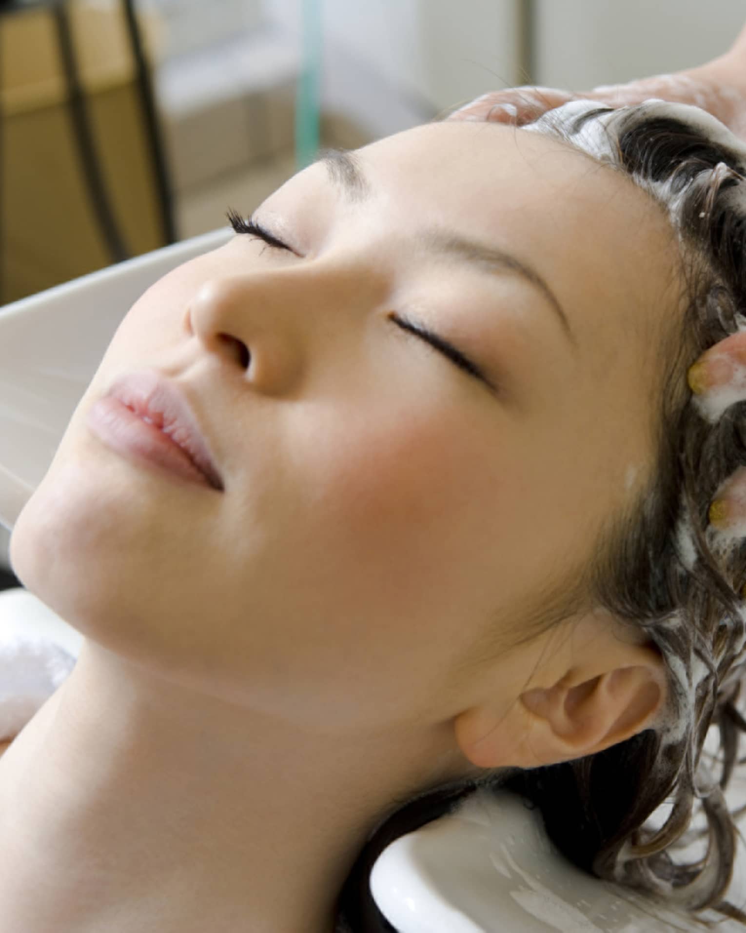 Woman closes eyes and leans back against sink as hands massage shampoo into her scalp