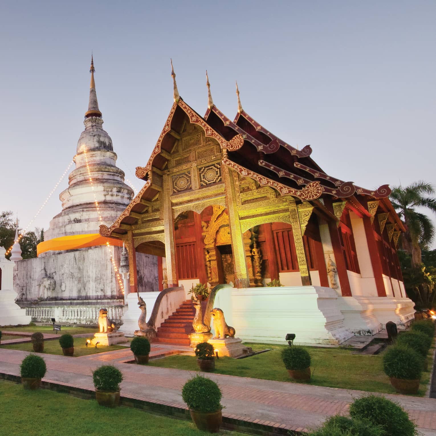 Two temples with gabled roofs and intricate carvings uplit in gold, between them a white domed building and stone monument.