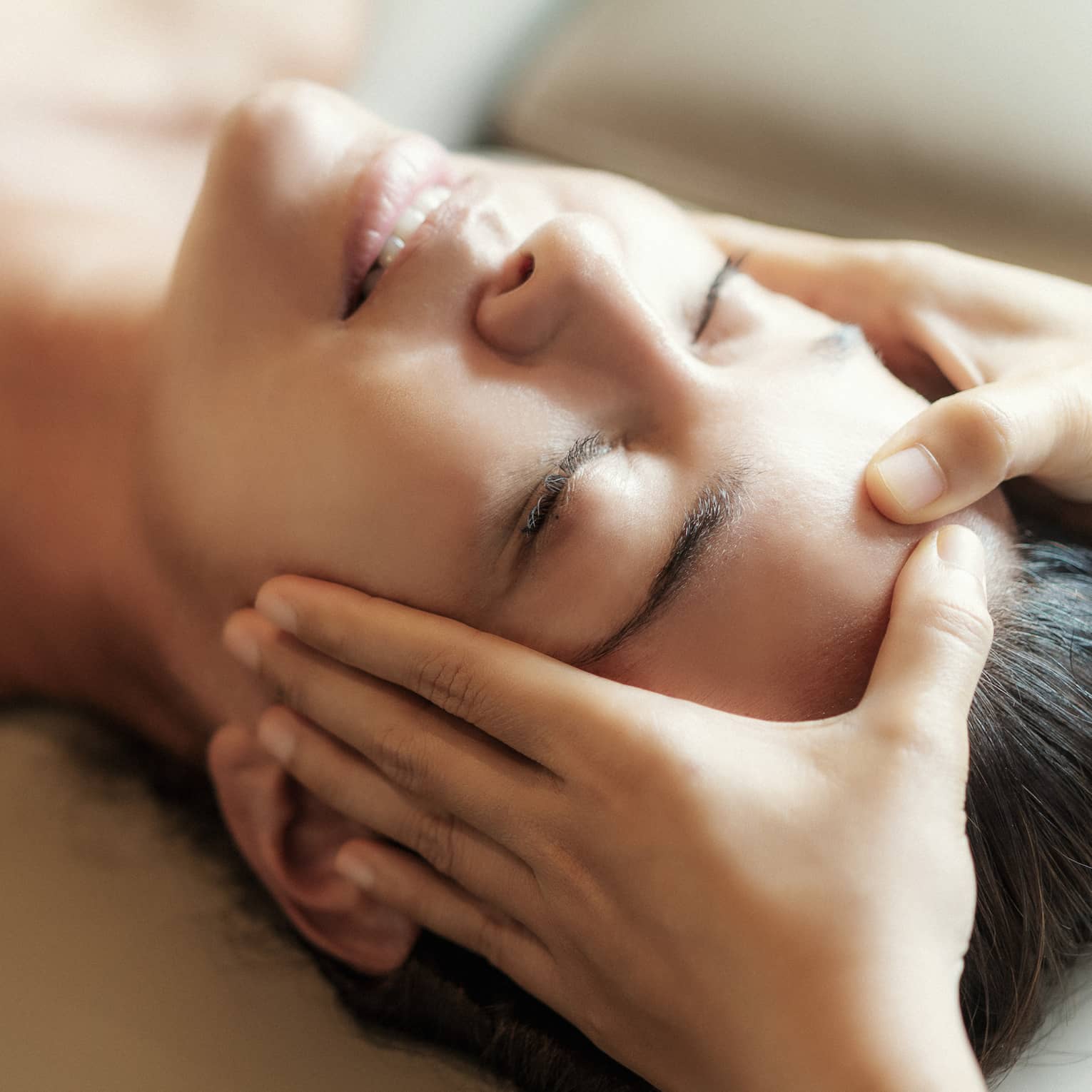 Spa facial, two hands rest on woman's forehead as she closes her eyes, lays under sheet on treatment table