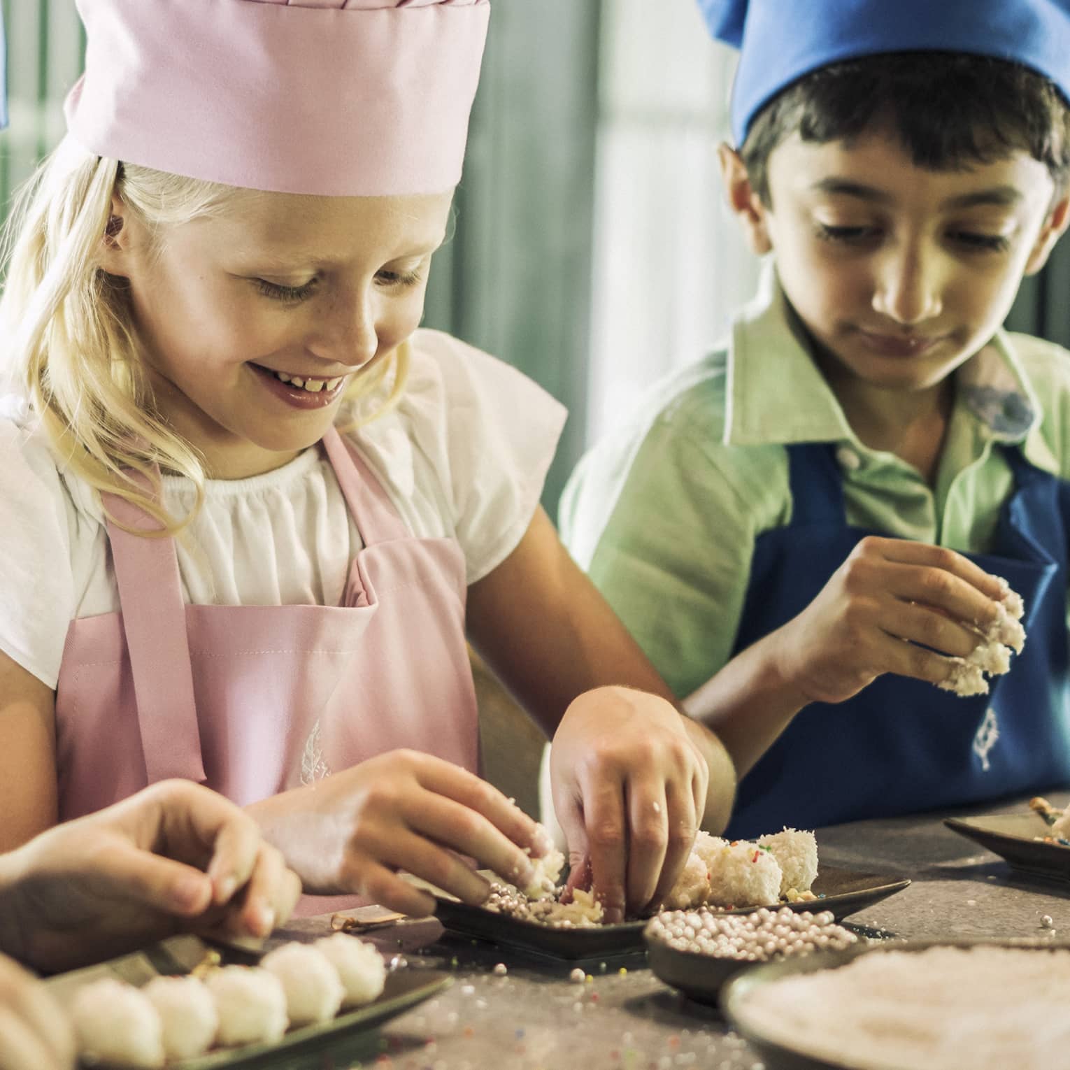 Three children in blue and pink aprons and chef hats roll pastries on small black trays