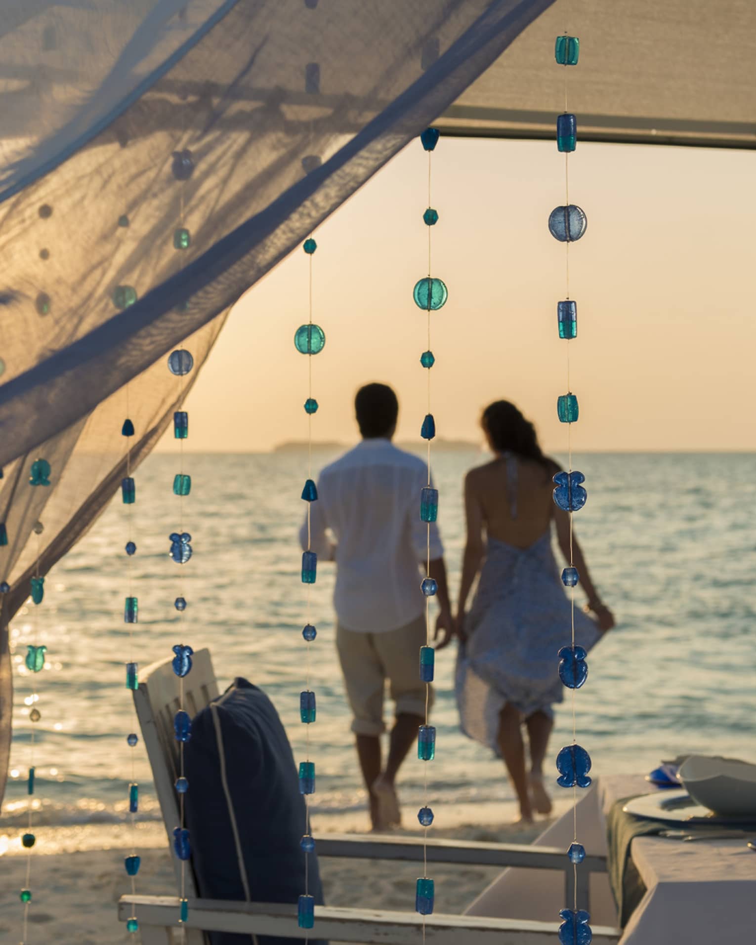 Sheer curtains blow in breeze, small blue beads hang from string, woman and man hold hands on beach in background
