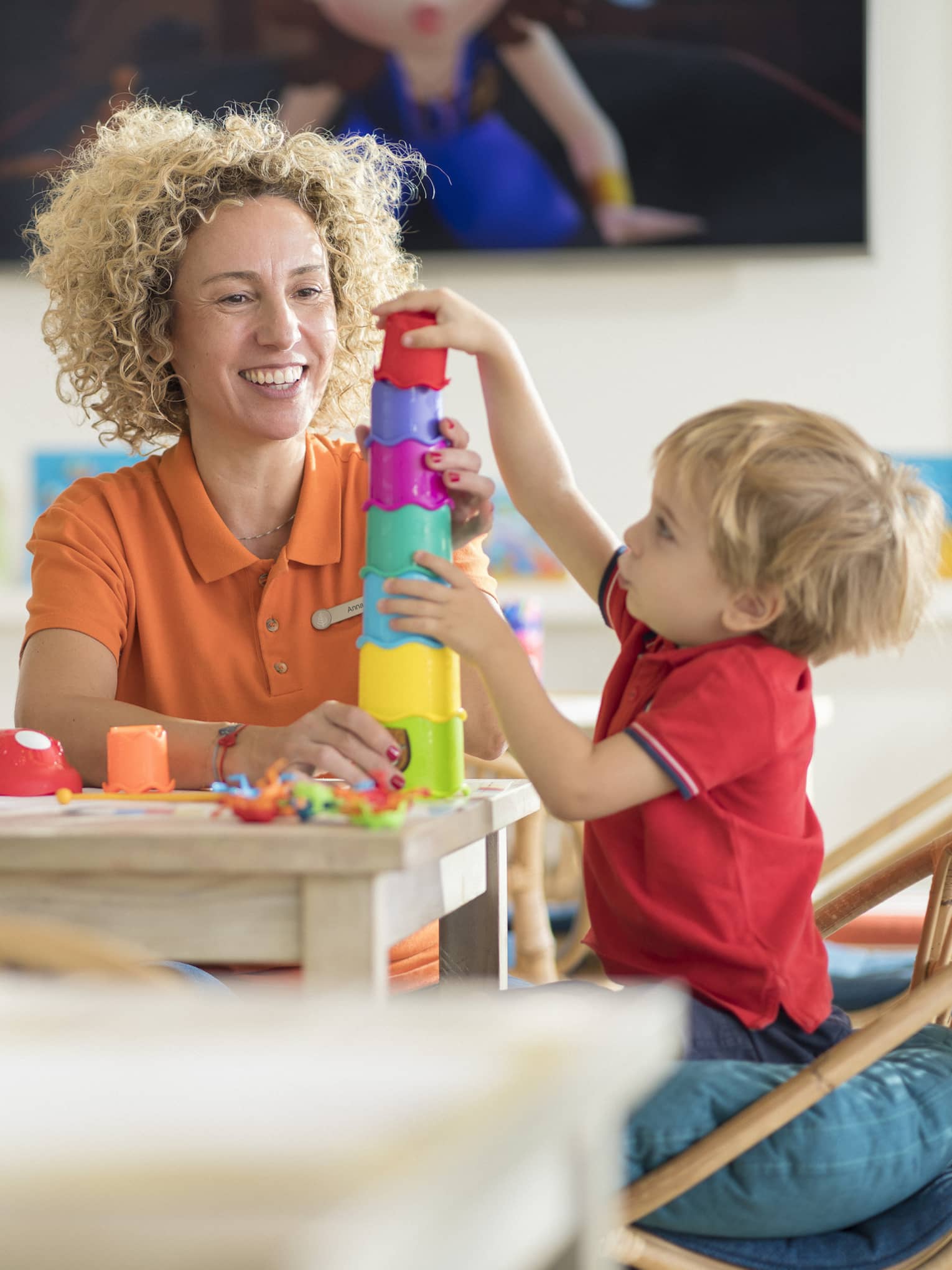 In a naturally lit and organized playroom, a daycare worker sits at a table with a young child playing with colourful blocks.