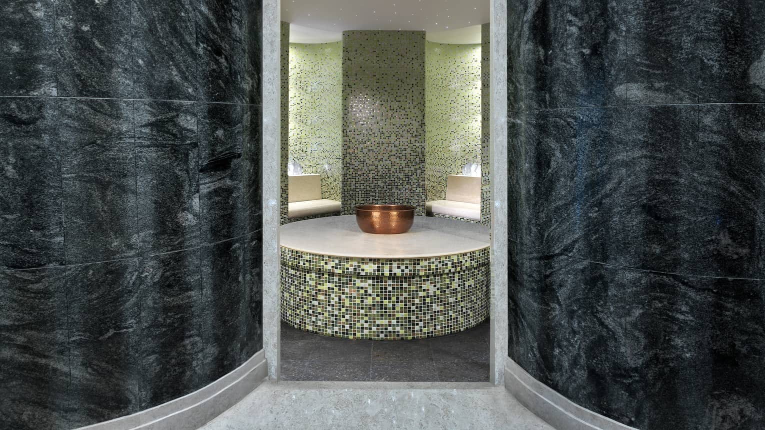 Hammam spa room with colourful tiles, curved black marble walls, copper bowl on round tile table