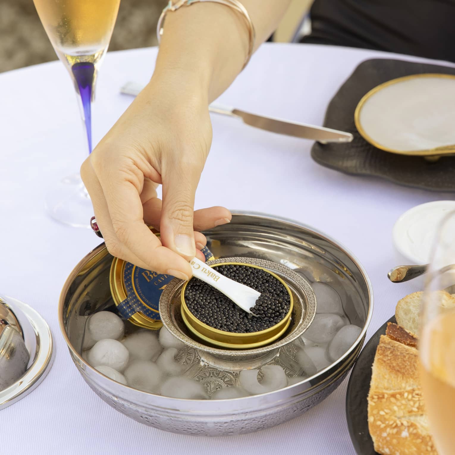 At a table set with champagne flutes, a diner holds buttered bread in one hand and spoons black caviar with the other.