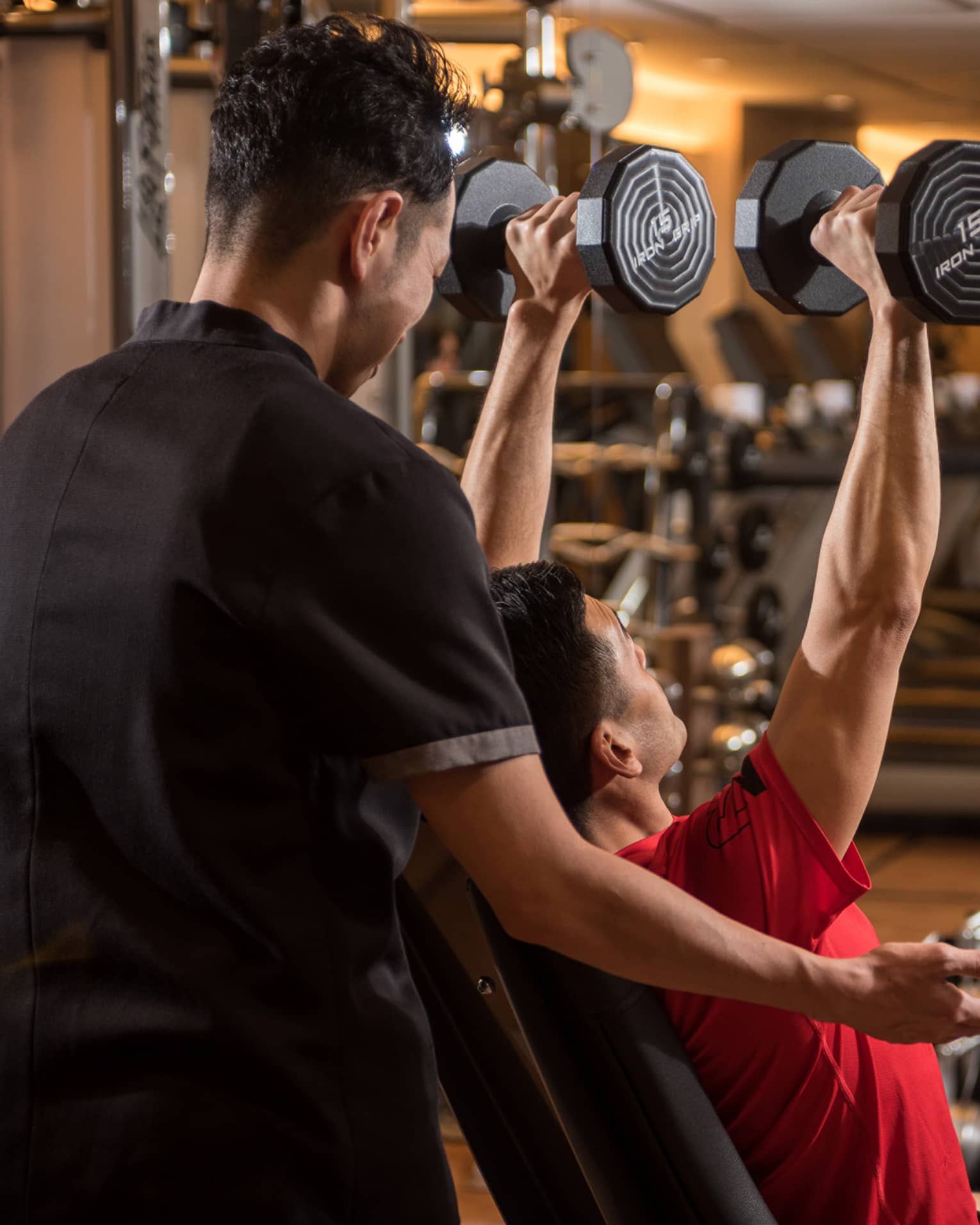 Man in chair in front of mirror lifts two 15 lb. weights above his head while personal trainer assists