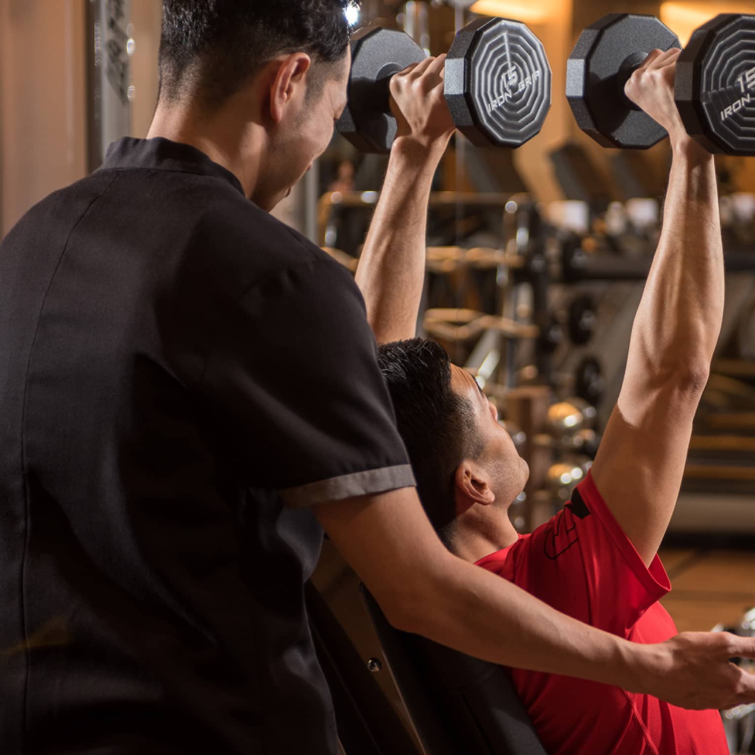 Man in chair in front of mirror lifts two 15 lb. weights above his head while personal trainer assists