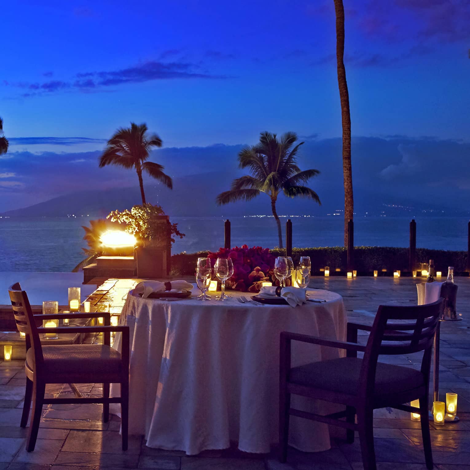 Romantic oceanfront dinner table setup at dusk with candles and palm trees