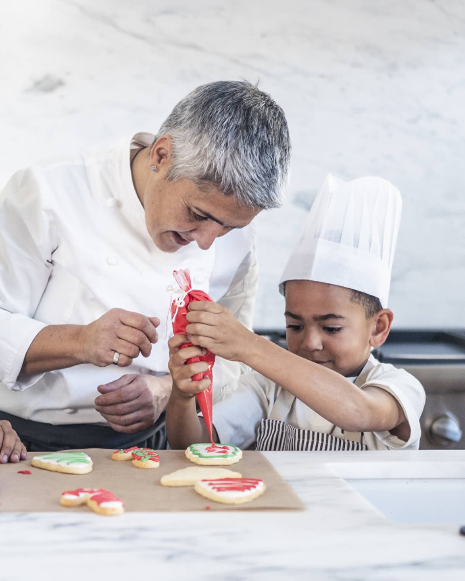 Two children in chef’s hats and aprons in a stark, white kitchen; a chef watches closely as one pipes icing onto a cookie.