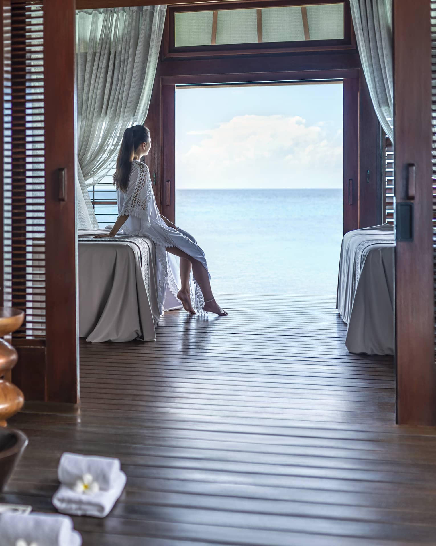 A woman leans on a massage bed on the far side of a spa treatment room and looks out through a large doorway onto the vast blue ocean