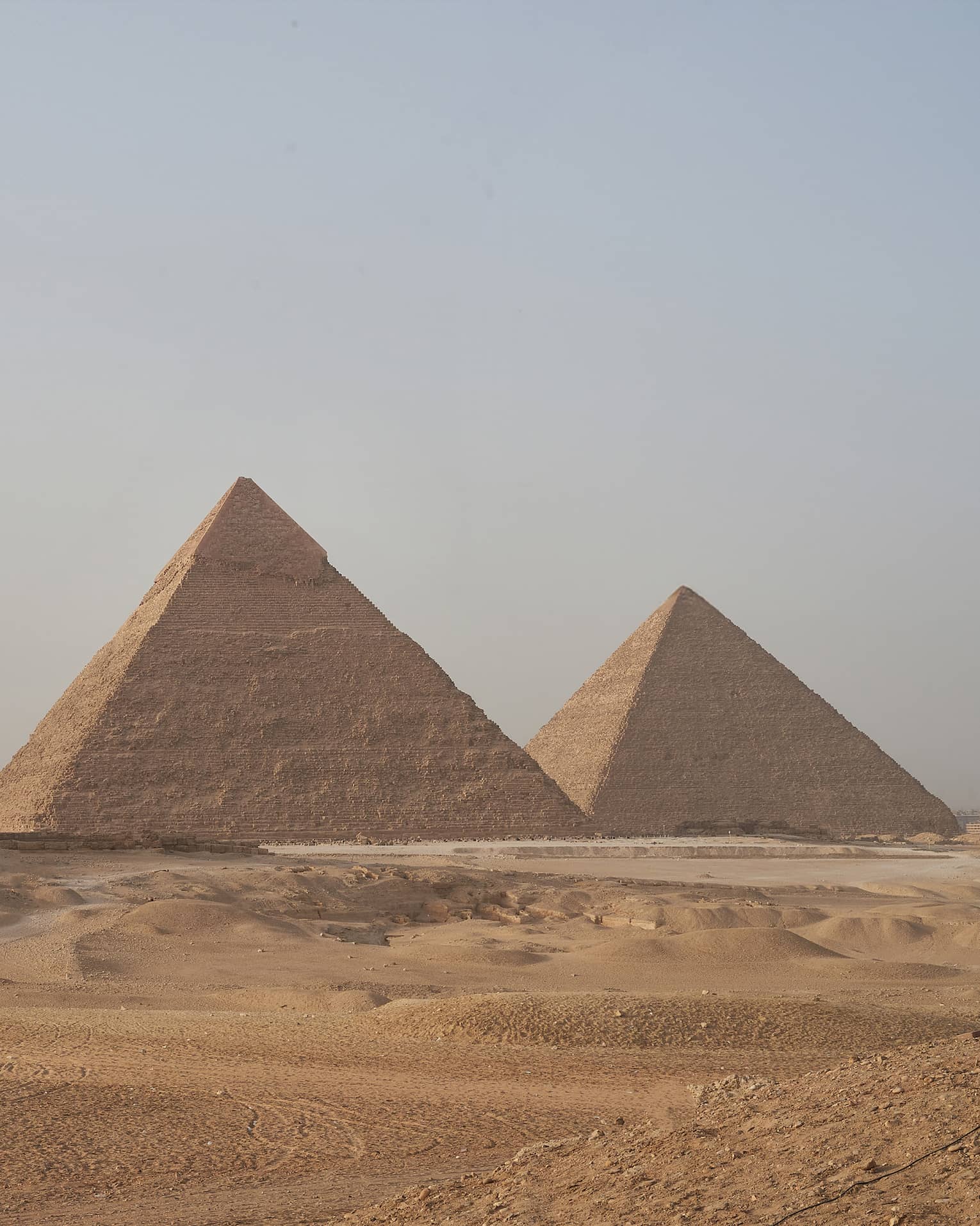 In the arid desert, a line of three towering pyramids dwarf two smaller pyramids before them, under a hazy blue sky.