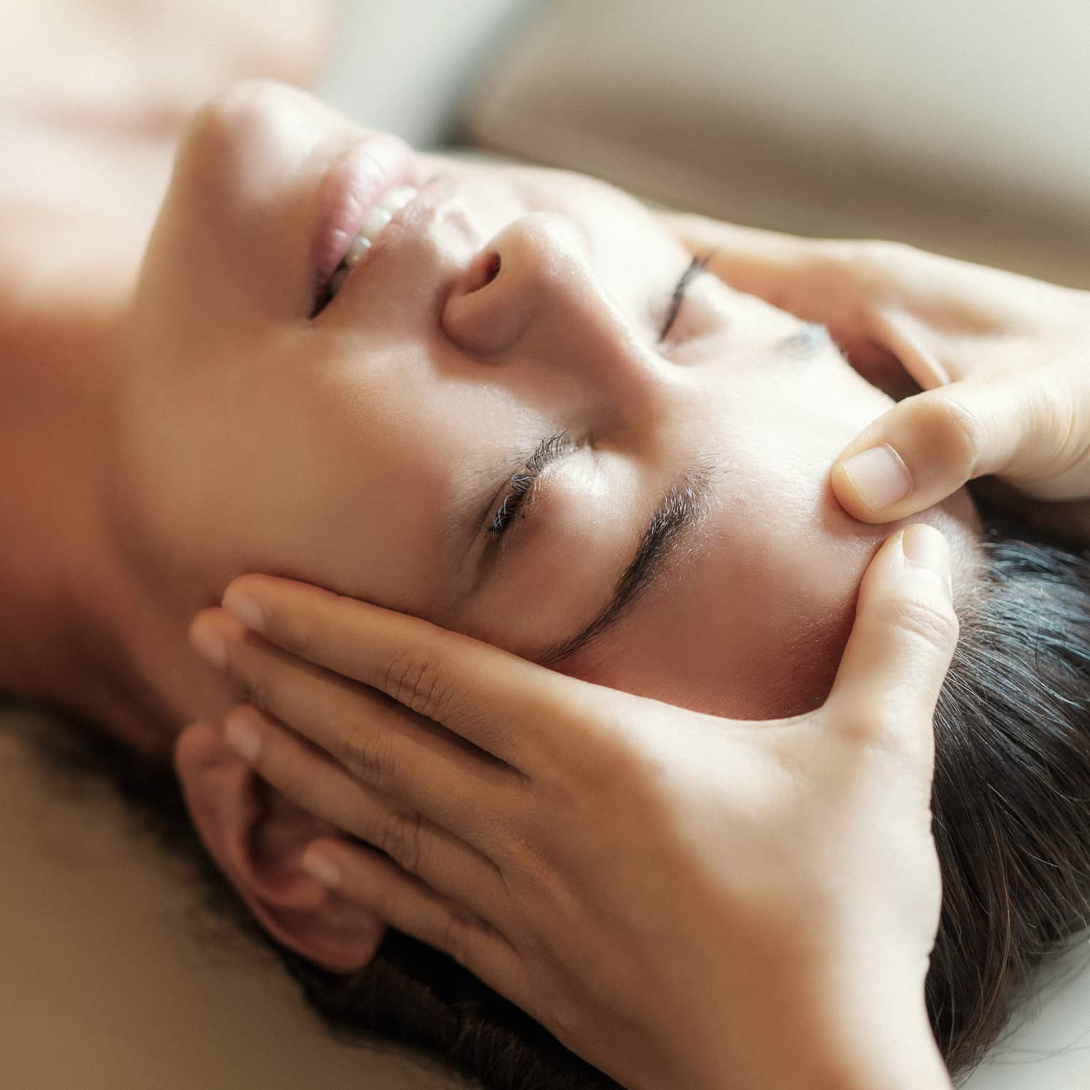 Spa facial, two hands rest on woman's forehead as she closes her eyes, lays under sheet on treatment table