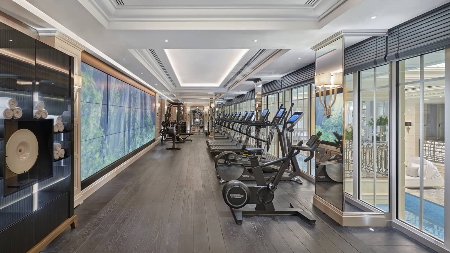 A row of cardio machines in an indoor gym, with windows looking out to indoor pool area