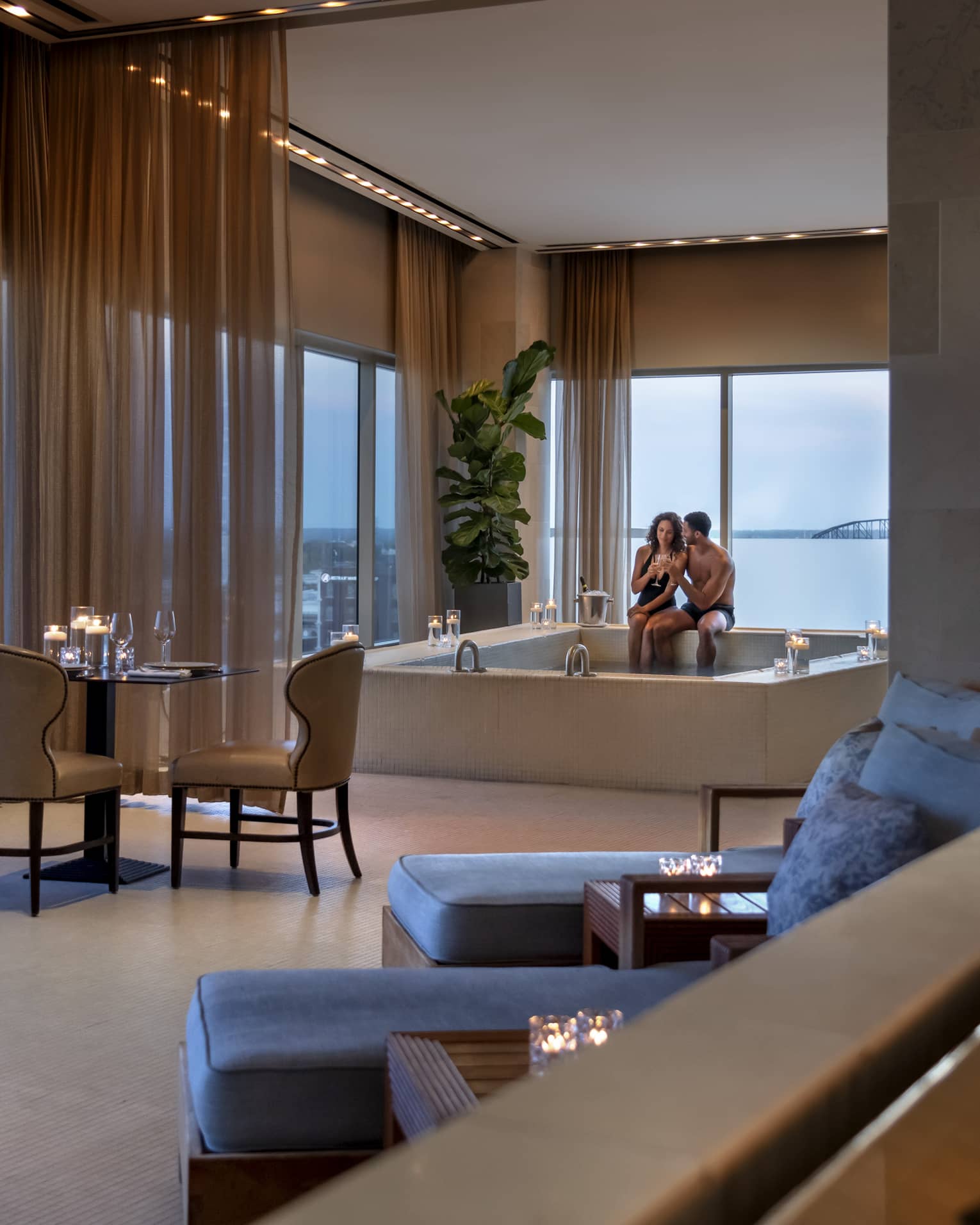 A couple enjoying a candlelit hot tub in a luxury hotel spa, with elegant seating, soft lighting and large windows offering a view of a bridge in the distance