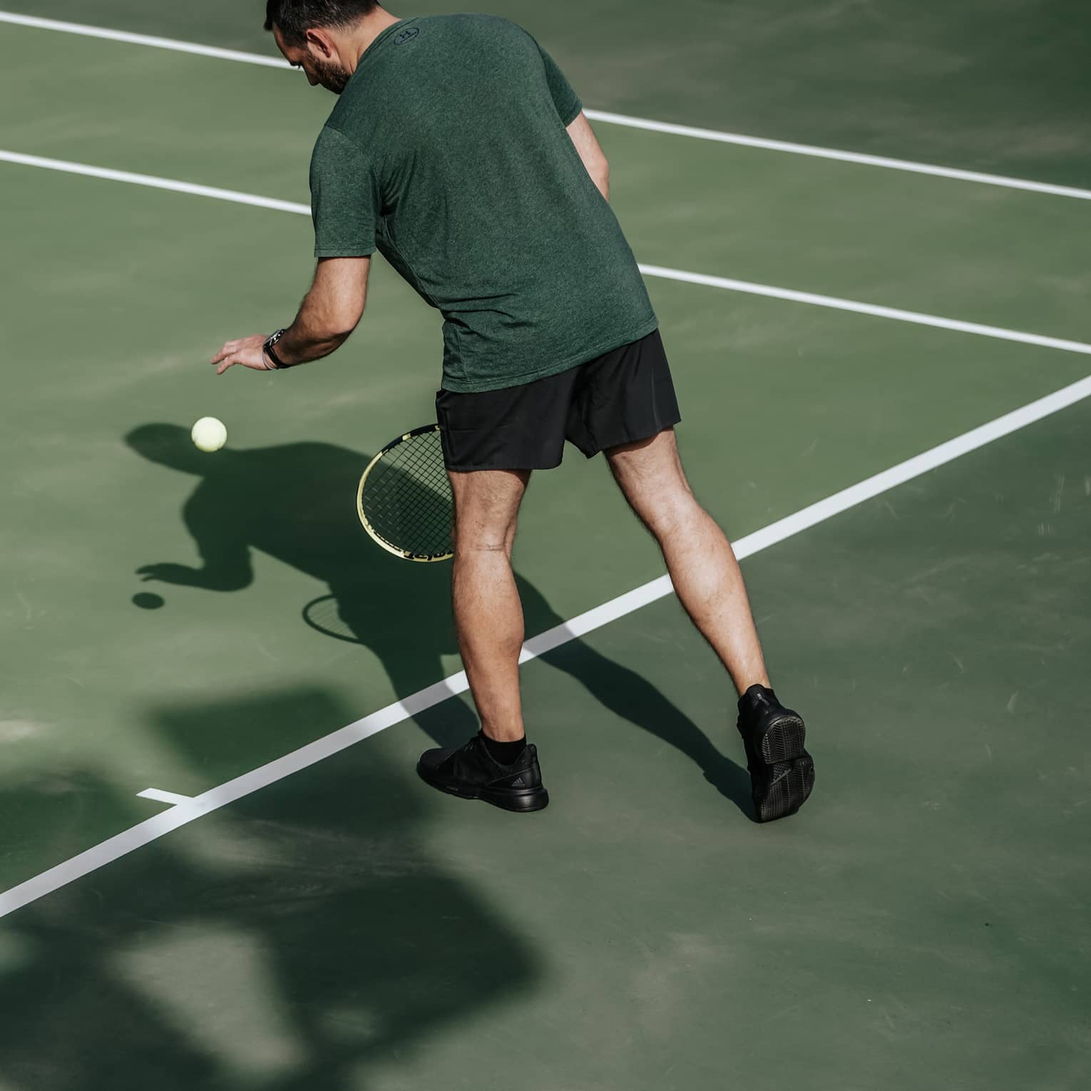 Man on tennis court bouncing tennis ball with left hand, preparing to serve