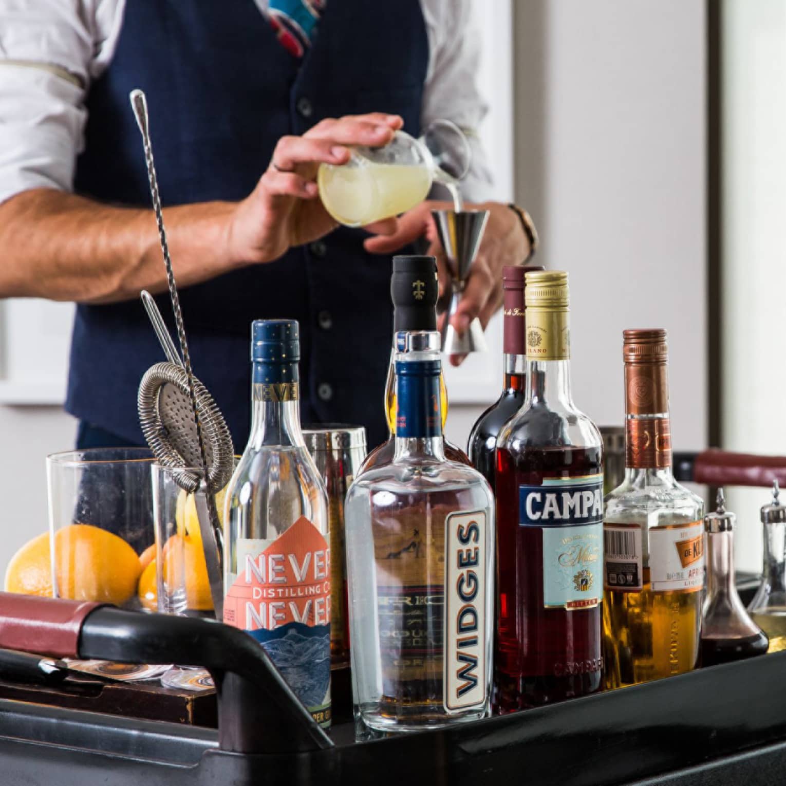 Bartender pours a cocktail at in room cocktail cart with assortment of liquor bottles