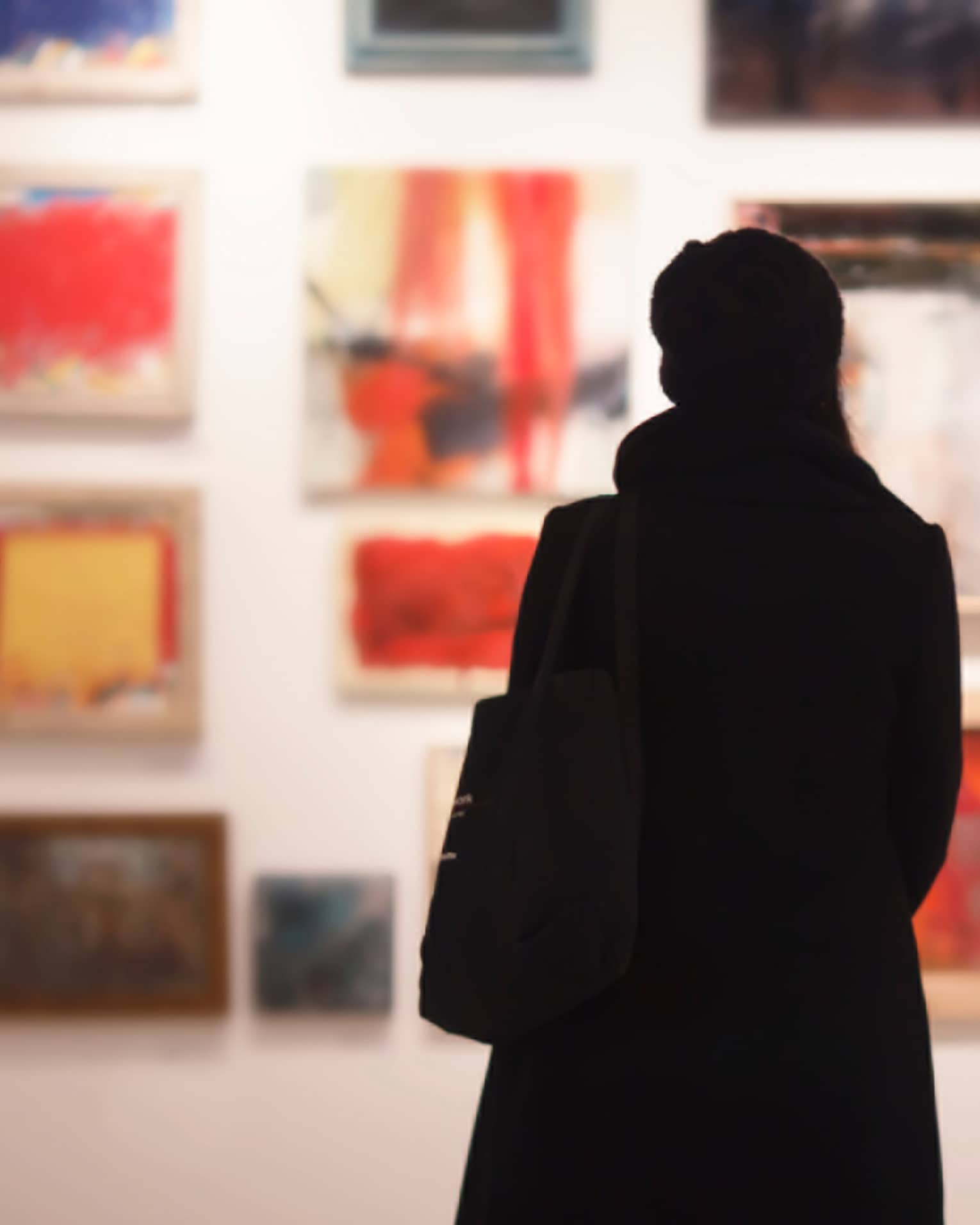 Silhouette of back of woman with hat bag looking at gallery wall with colourful art