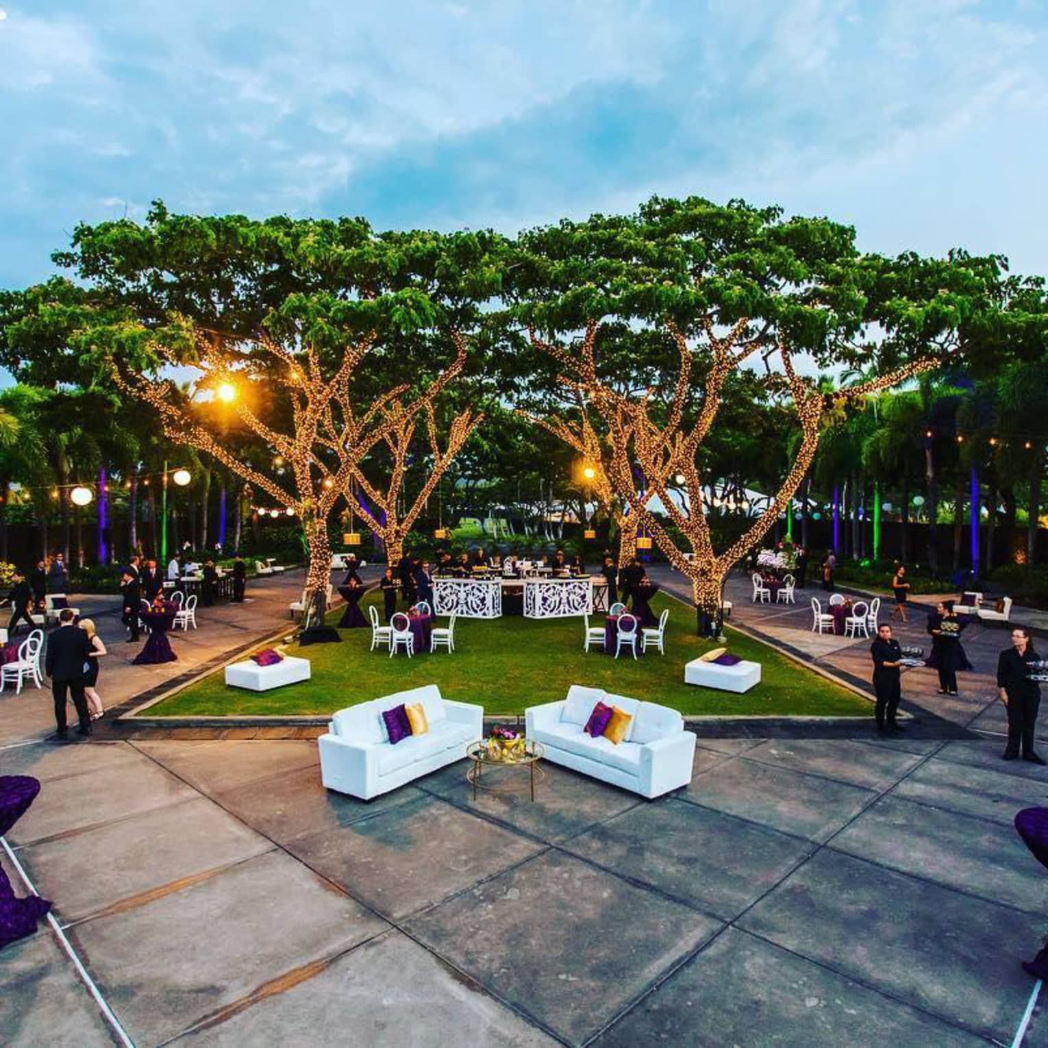 Guests and servers stand in a lit courtyard filled with tables, chairs and plush white sofas
