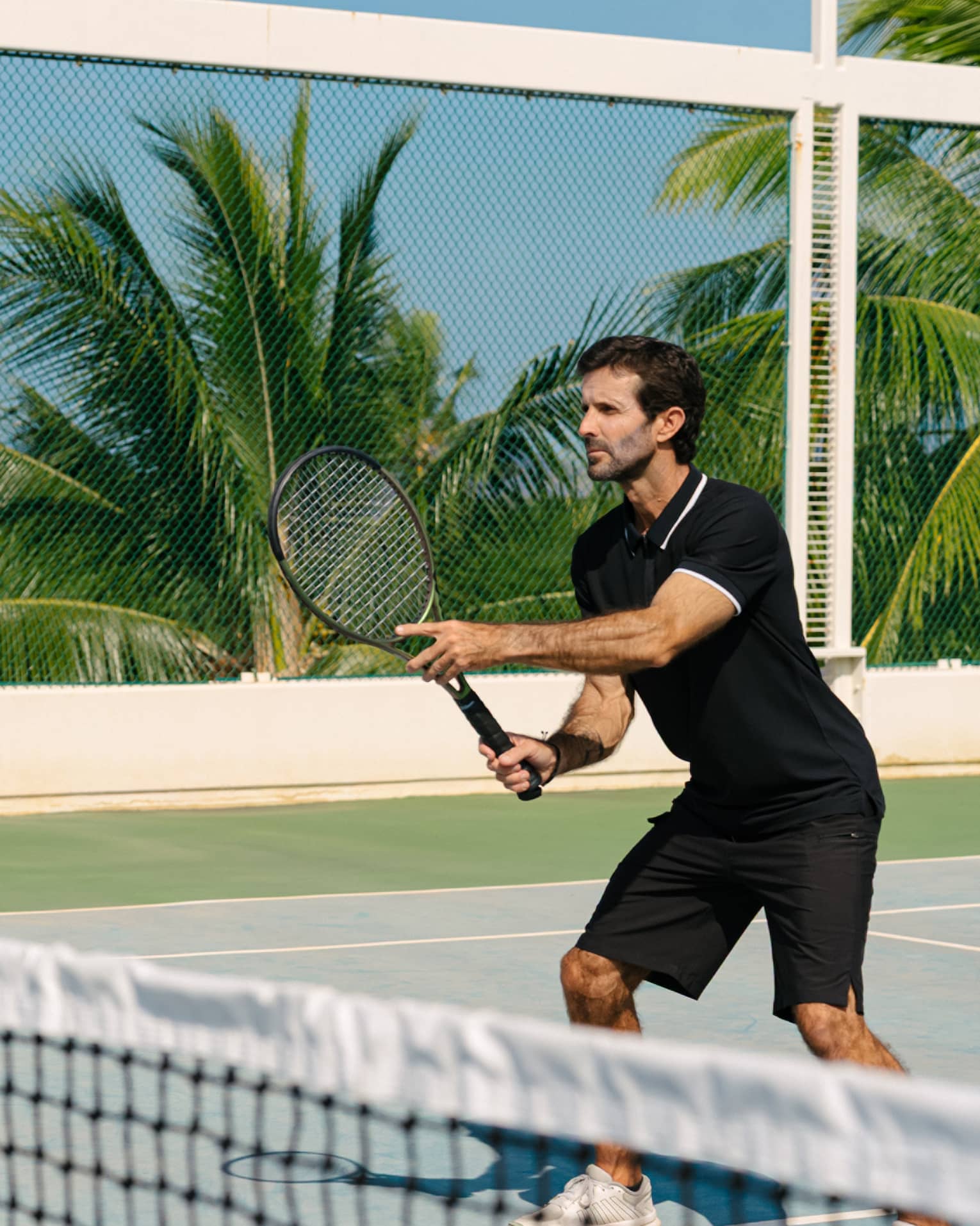 A man wearing a black polo and shorts plays tennis in tropical resort location