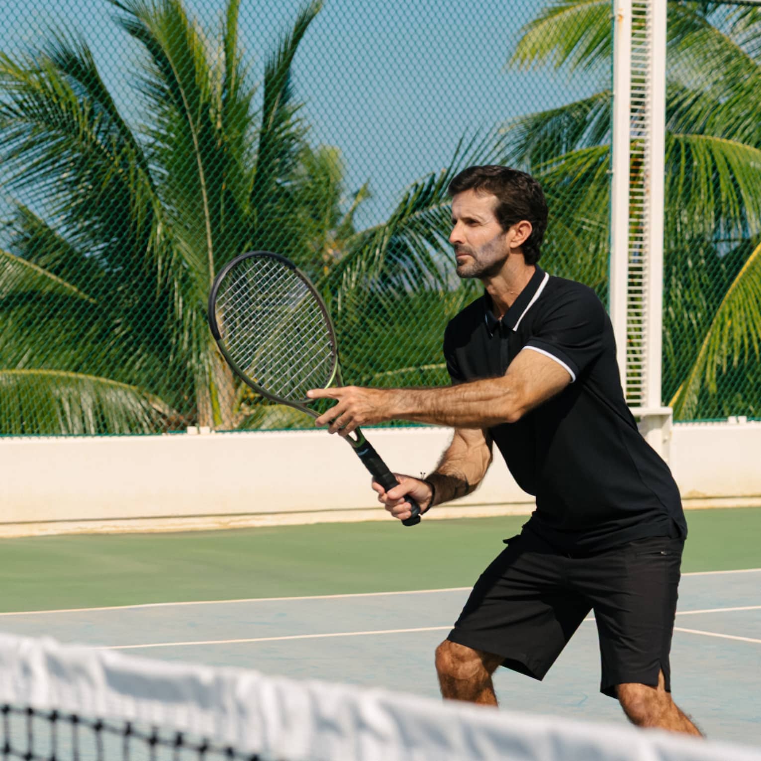 A man wearing a black polo and shorts plays tennis in tropical resort location