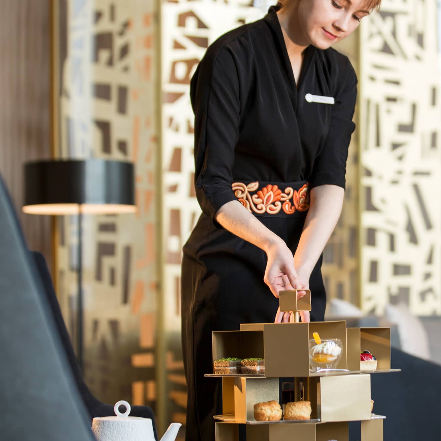 Server holds geometric gold tray with Afternoon Tea small gourmet desserts on lounge table near modern grey chair, pillow