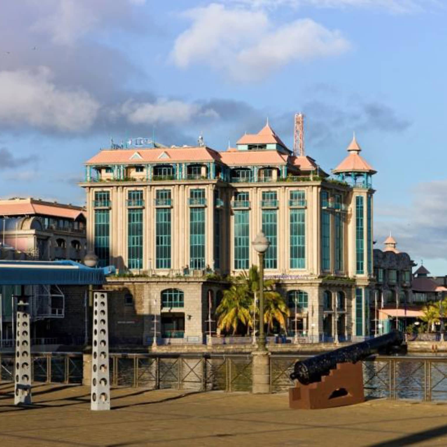 An old cannon in the foreground borders a channel, with ornate Colonial-style buildings across the channel on the shoreline.
