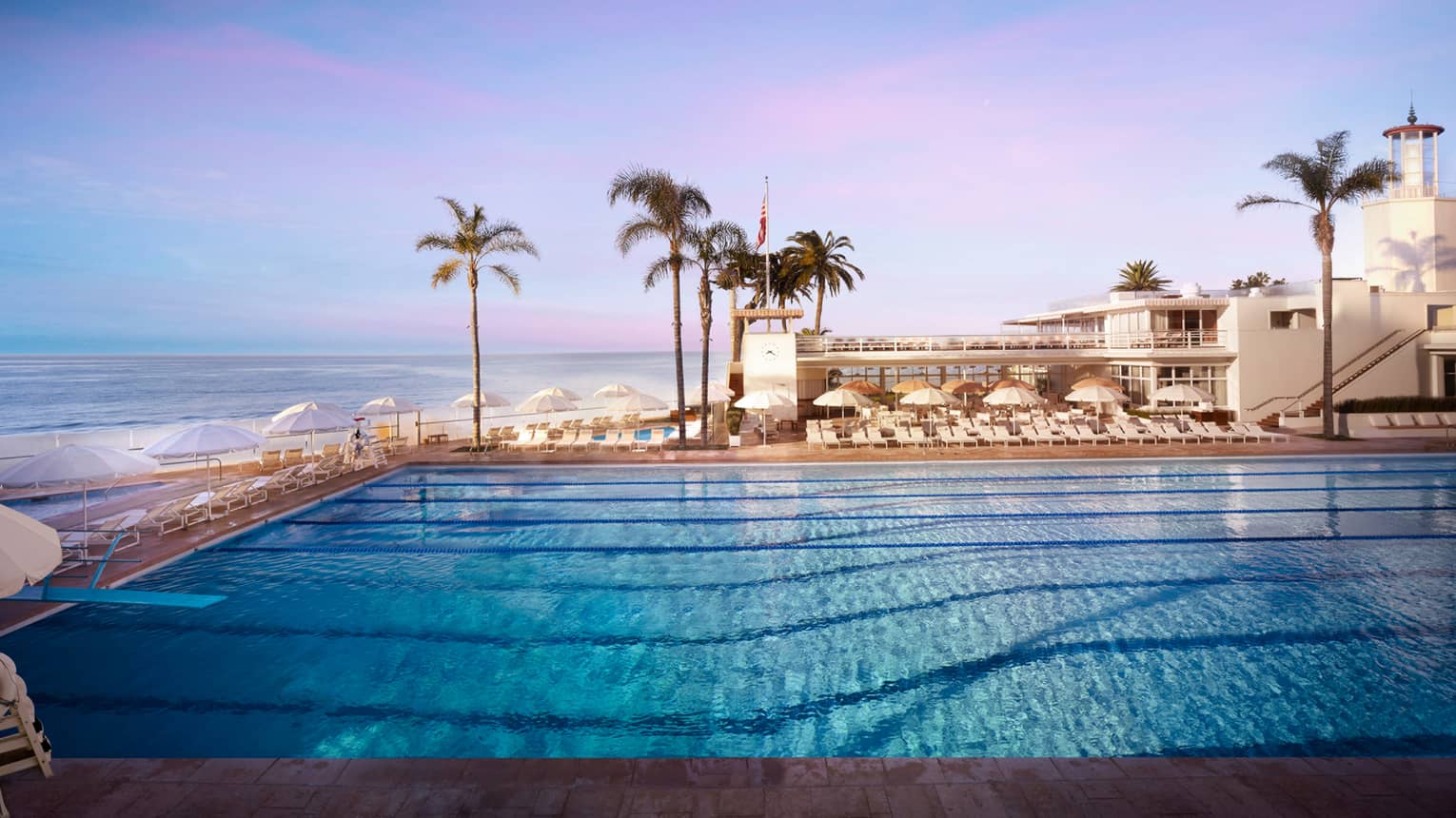 White pool chairs, umbrellas line Olympic-size outdoor swimming pool, ocean in background