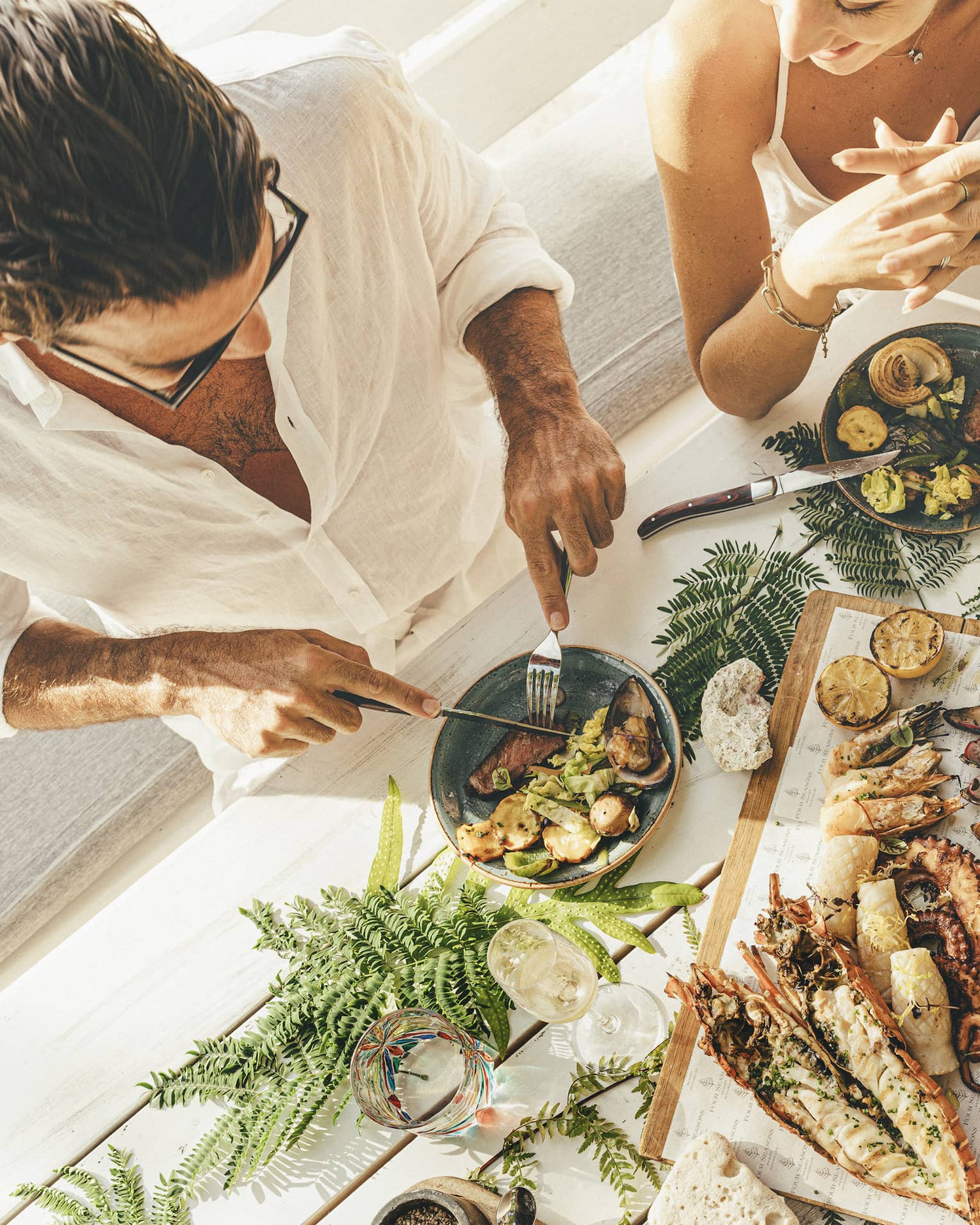 A couple at a table of seafood canapés, grilled meat, corn and champagne flutes, adorned with palm fronds and red hibiscus.