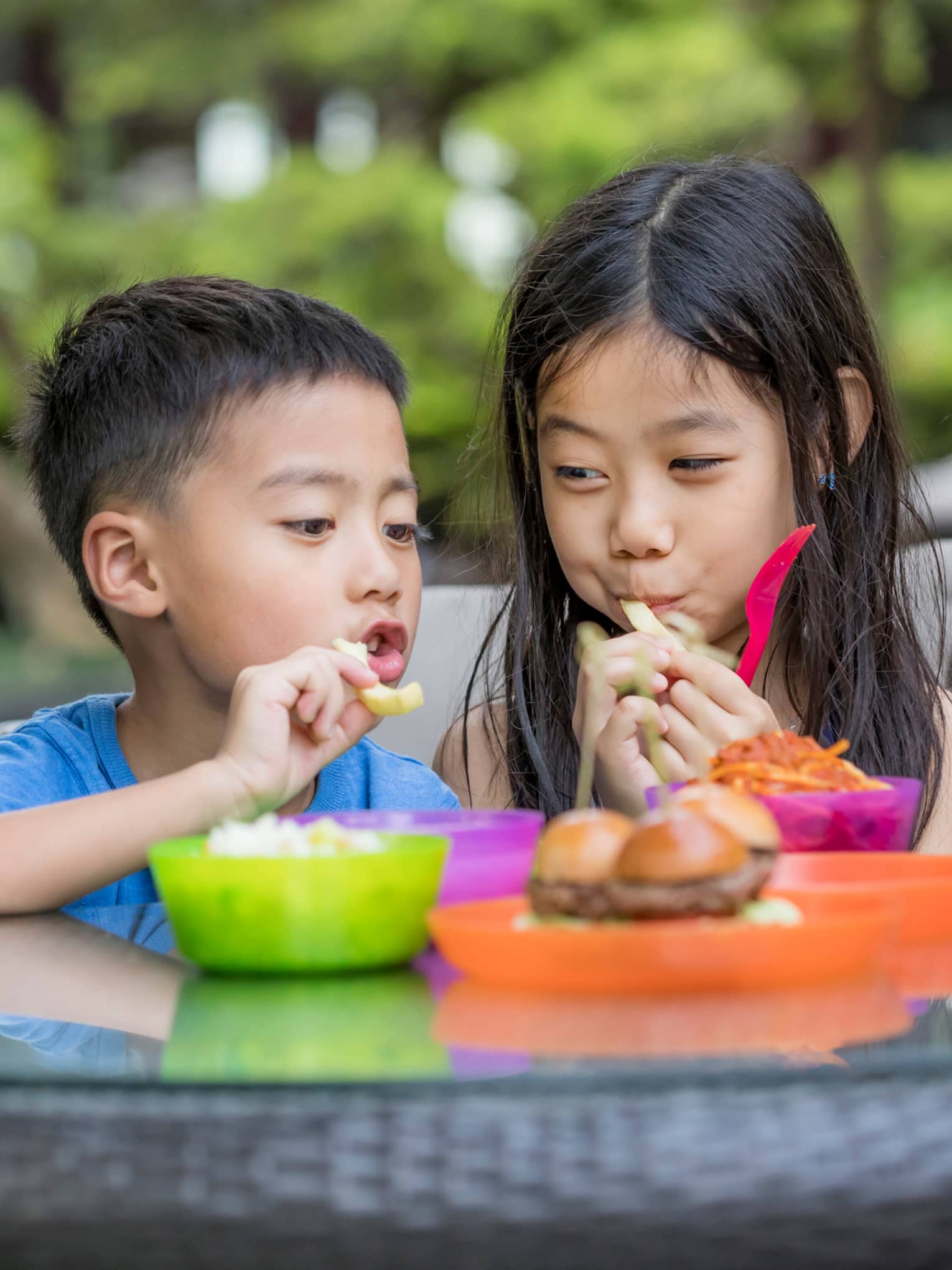 Children enjoying a variety of dishes in colourful plastic dinnerware on a glass-top table on a patio surrounded by greenery.