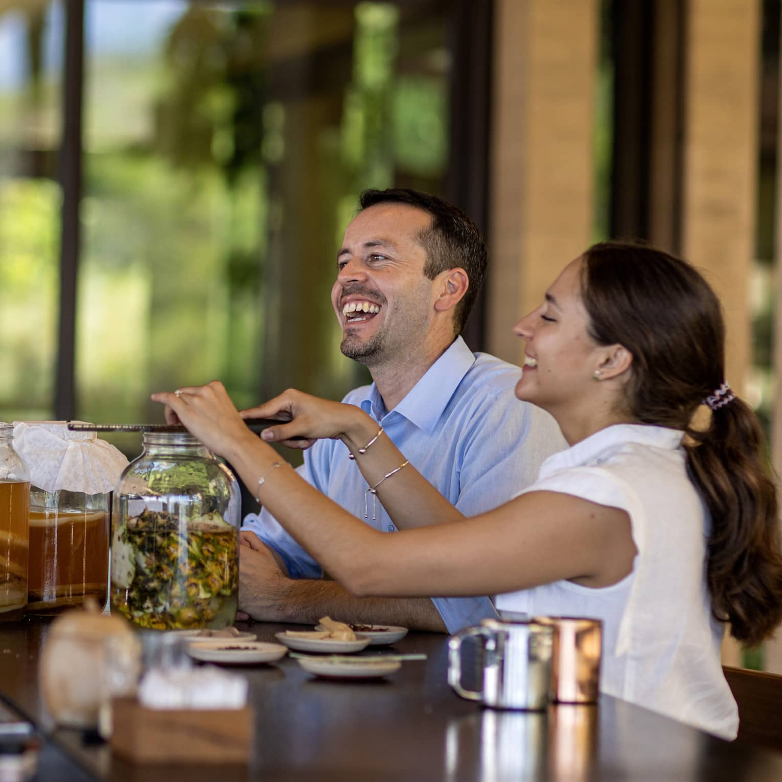 Three guests gather at a bar, one grates an ingredient into a large fermentation jar aside two other jars of amber liquid.