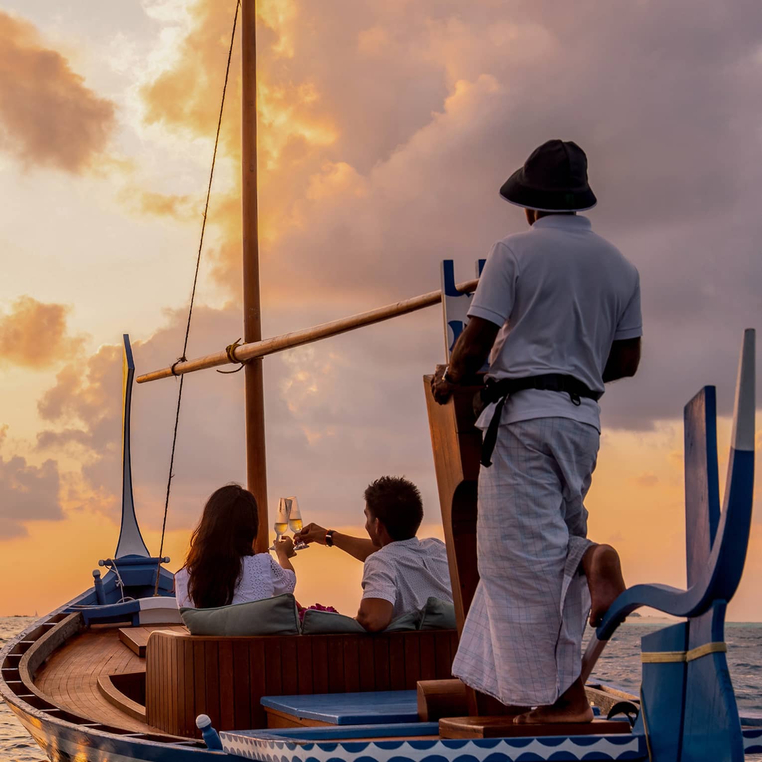 Rear view of two people seated in a dhoni boat, toasting with champagne, the boat handler steering into the golden sunset.