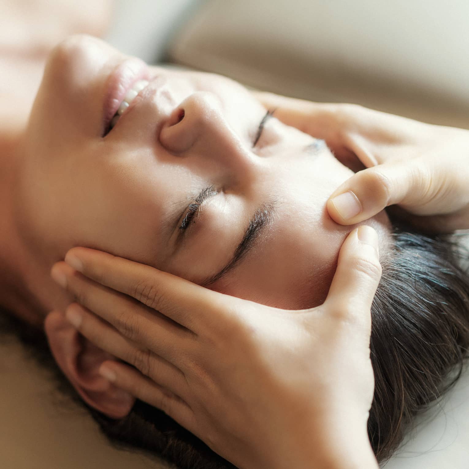 Spa facial, two hands rest on woman's forehead as she closes her eyes, lays under sheet on treatment table