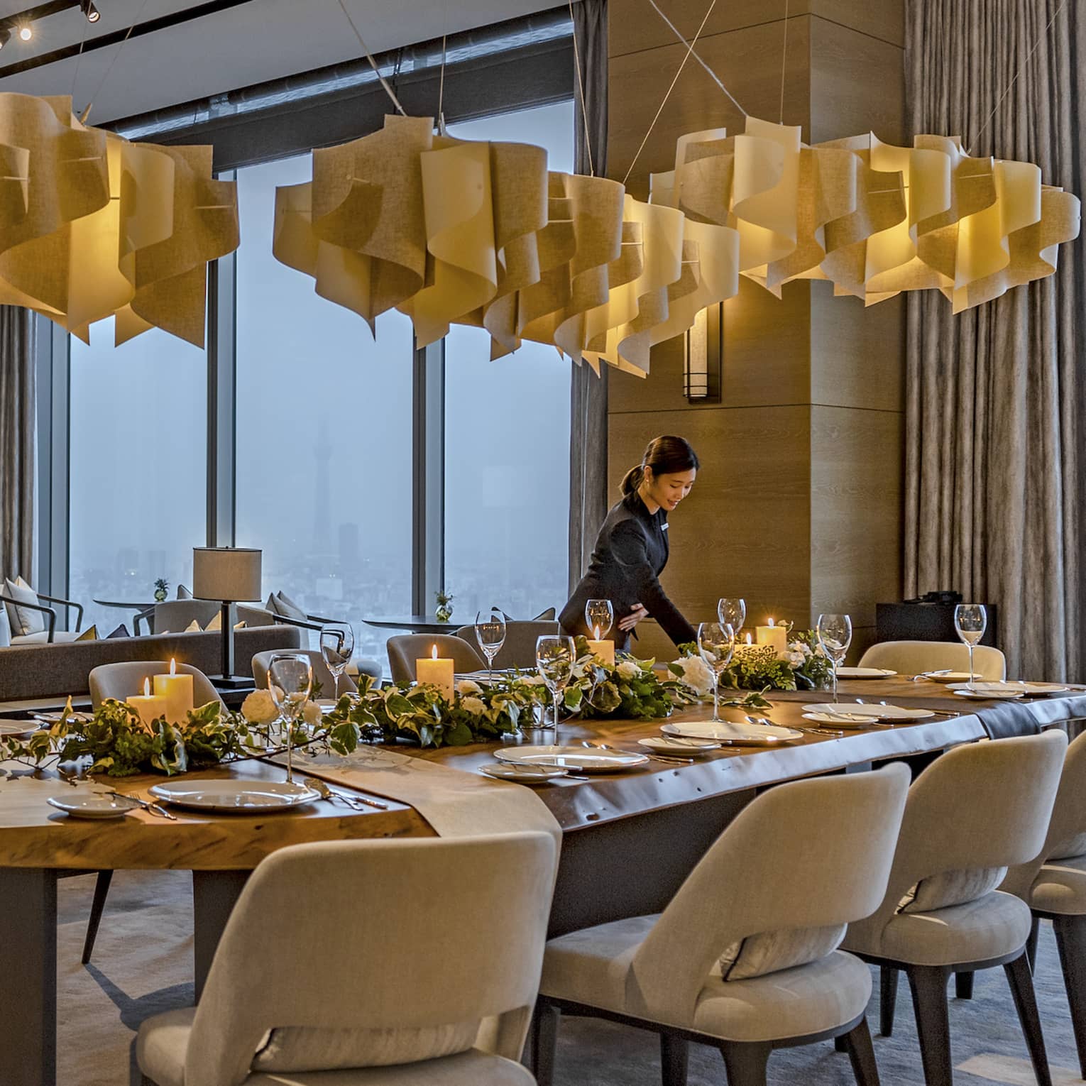 A candlelight dinner is set at a long, oval table with plush seating for twelve under pendant lamps made of swirling wood.