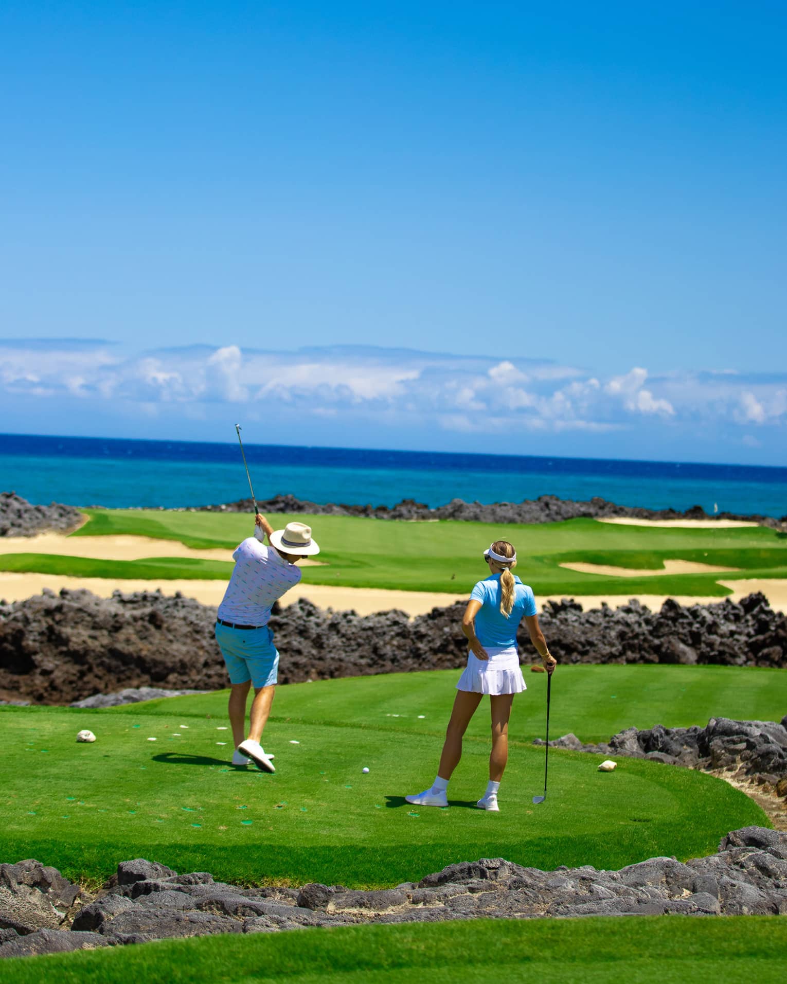Two people playing golf on green fairways with black lava rock and the ocean in the background.
