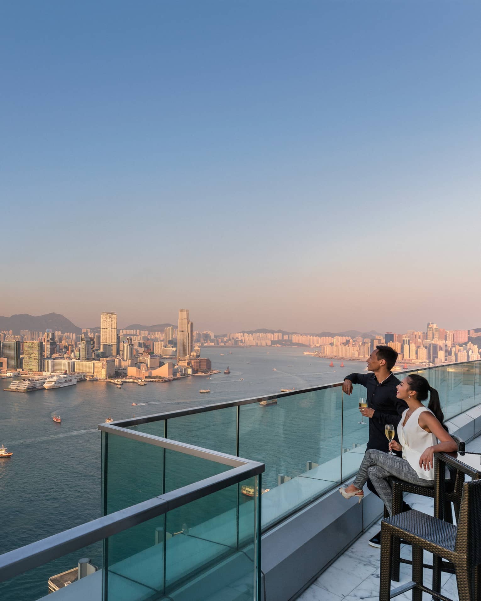 Couple on glass-walled terrace holding glasses of white wine, smiling and looking out over Hong Kong's Victoria Harbour at sunset