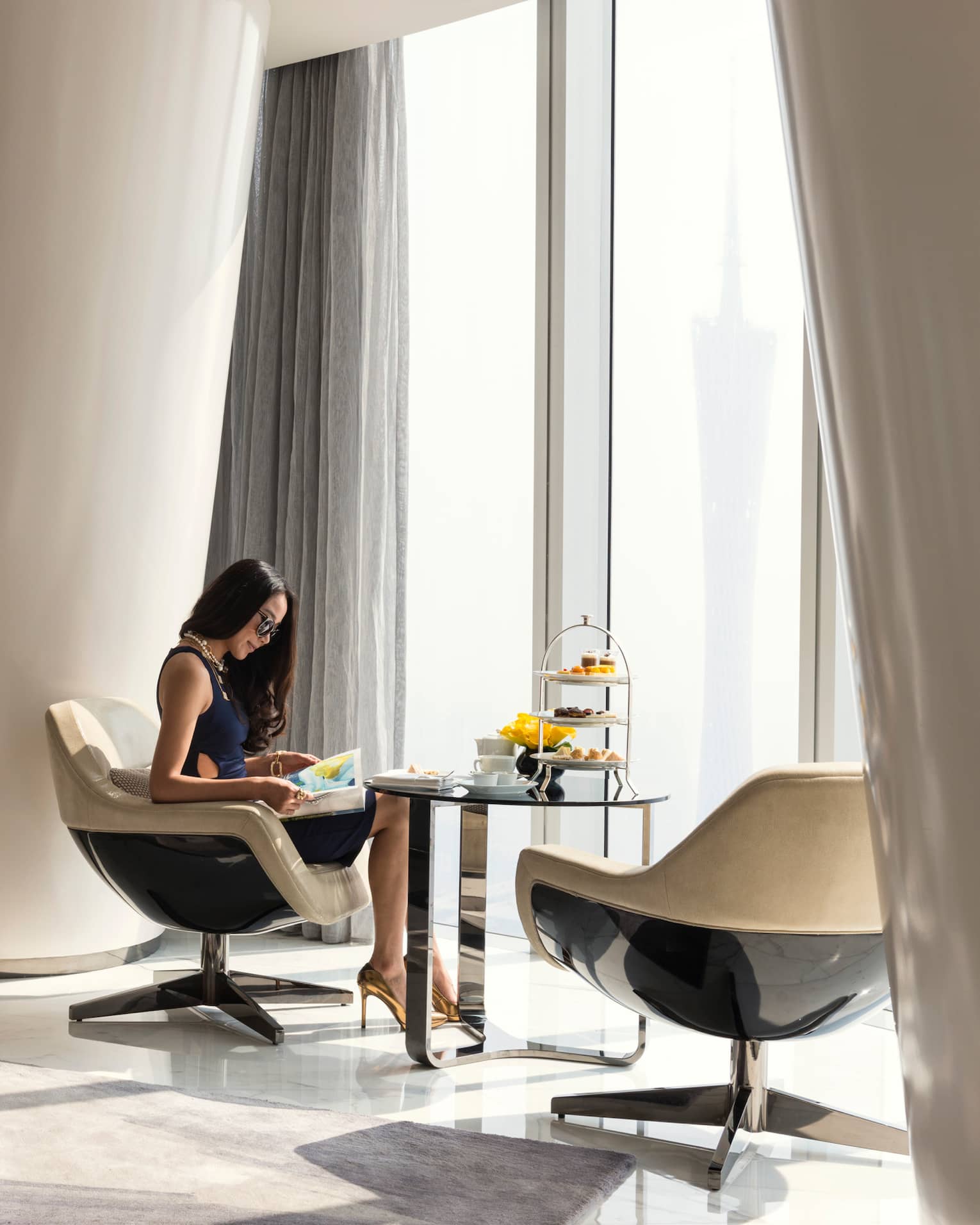 A guest sits in a neutral-toned room with a panoramic view and curved, modern design, a tiered tray of colourful fare nearby.