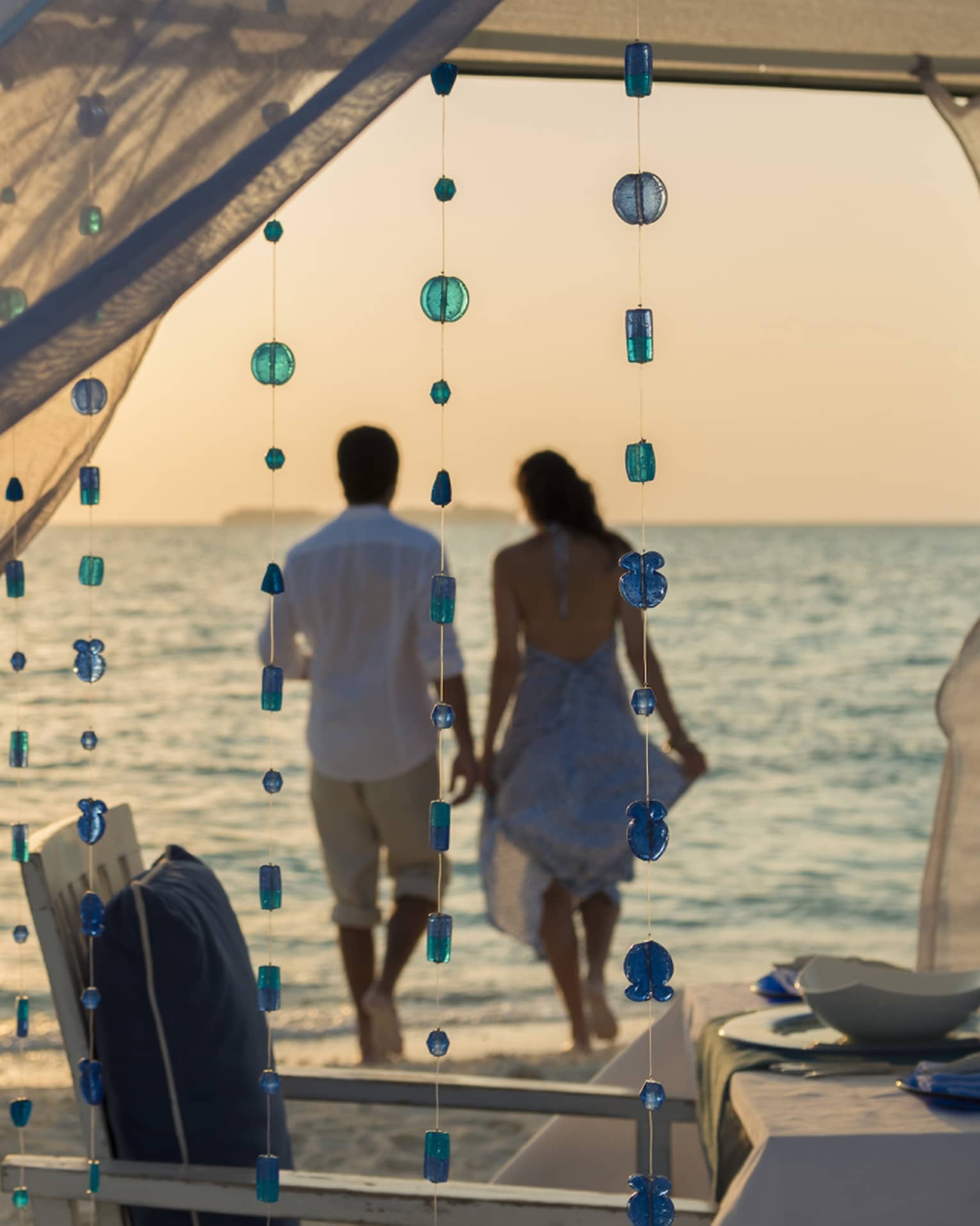 Sheer curtains blow in breeze, small blue beads hang from string, woman and man hold hands on beach in background