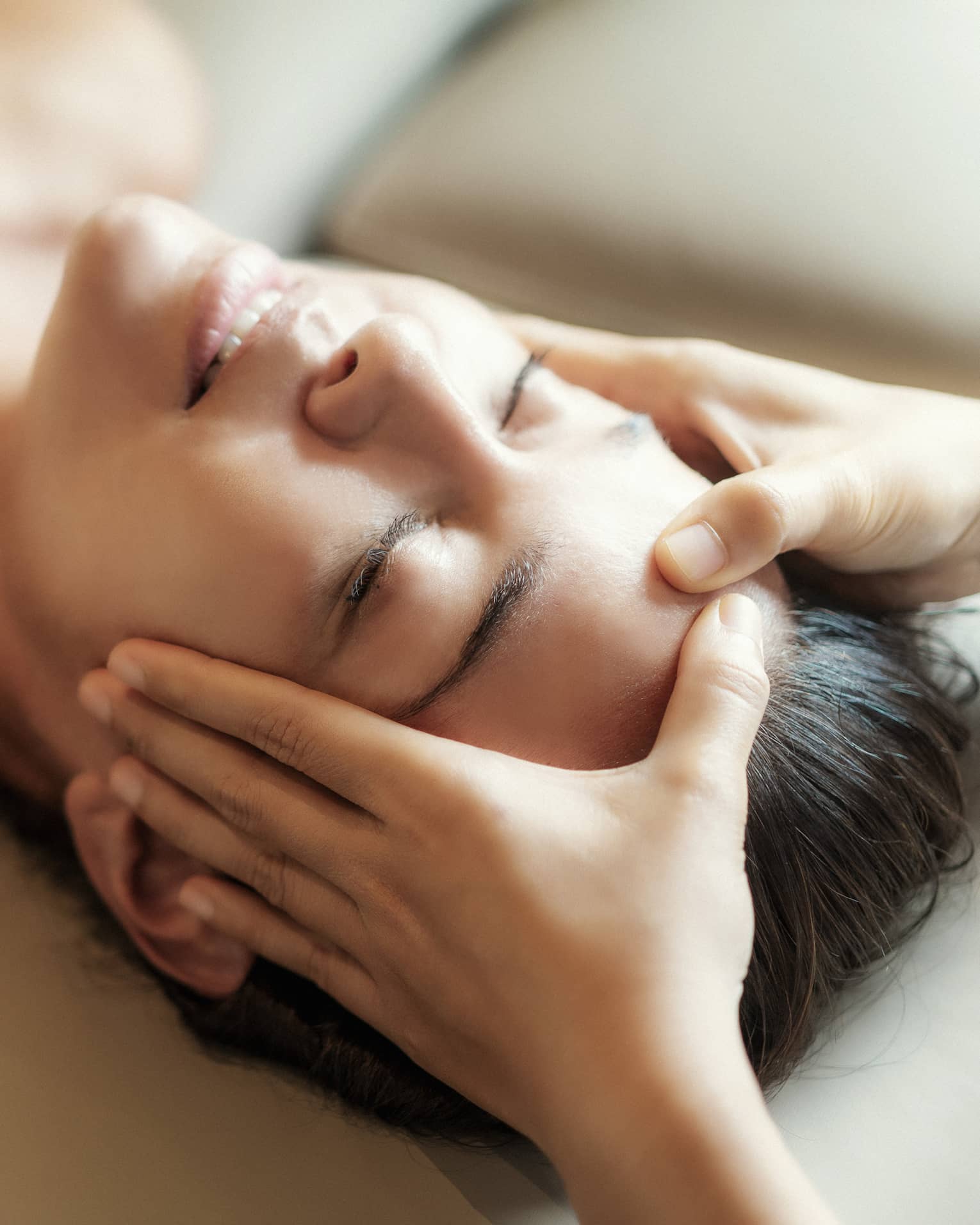 Spa facial, two hands rest on woman's forehead as she closes her eyes, lays under sheet on treatment table