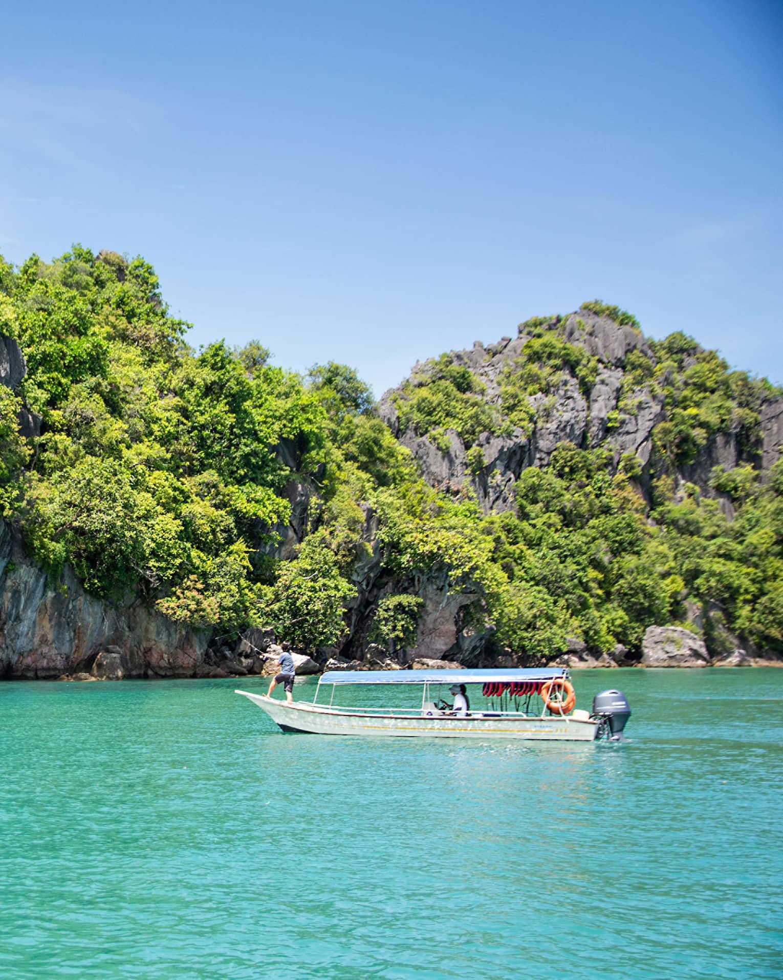 Guest fishing on a boat in turquoise water floating past a forested island with a tall craggy rock face under a blue sky.