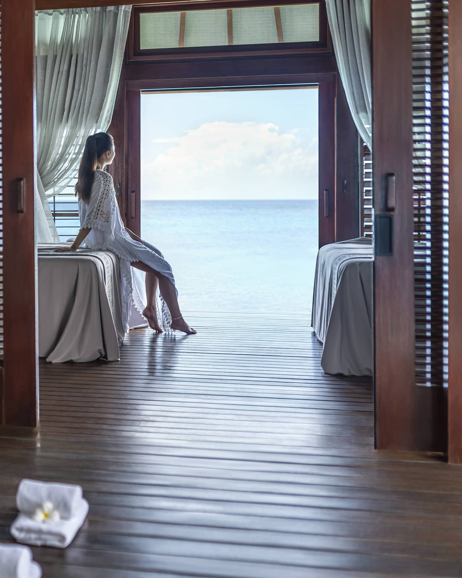 A woman leans on a massage bed on the far side of a spa treatment room and looks out through a large doorway onto the vast blue ocean