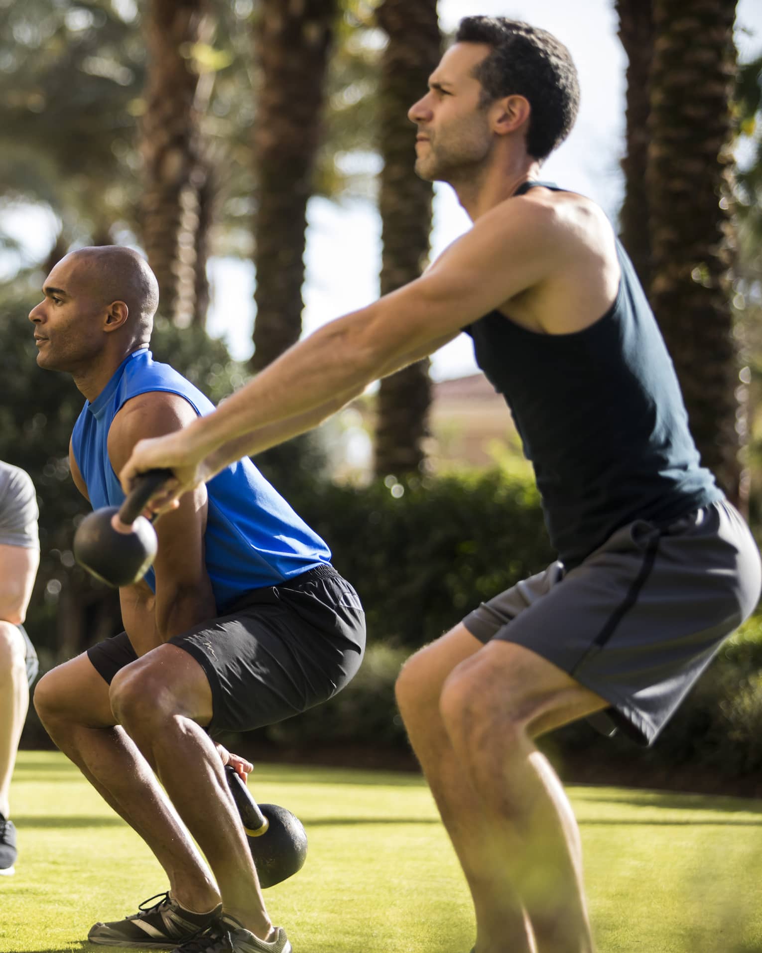 Four Seasons trainer watches as two men squat, lift kettlebell weights on lawn