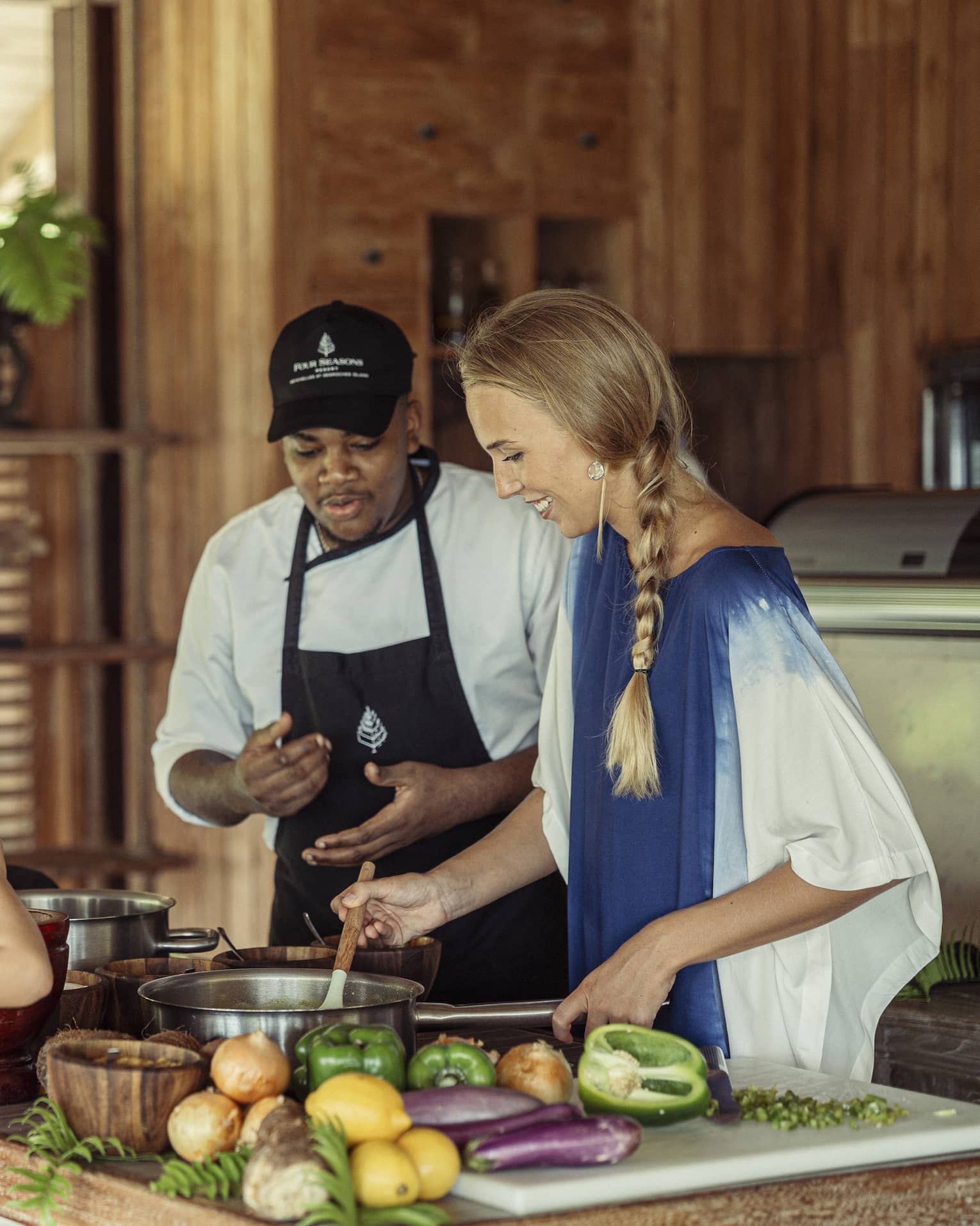 A chef instructs a guest stirring a pot, while a child uses a mortar and pestle across the counter as a third adult watches.