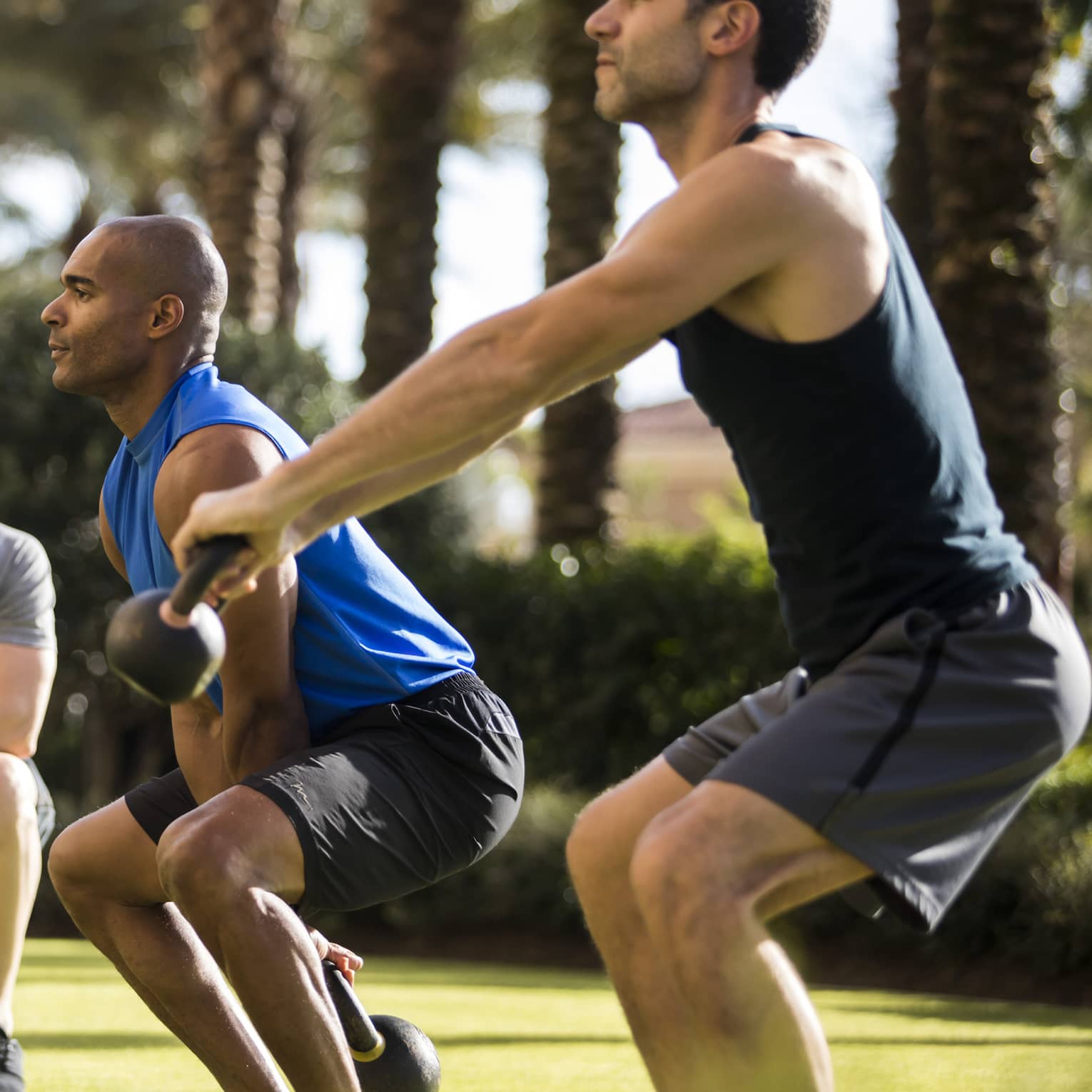 Four Seasons trainer watches as two men squat, lift kettlebell weights on lawn