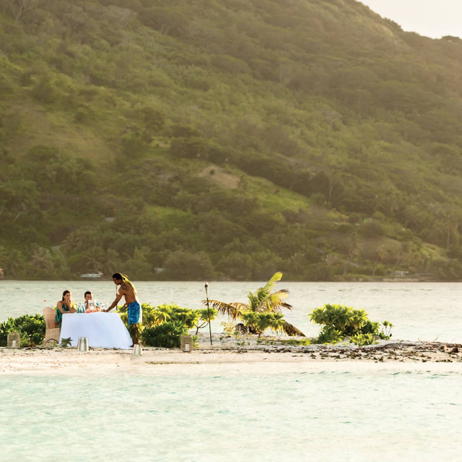 Small tropical island with palm trees, where two people are dining at a table by the water, with a mountain in the background
