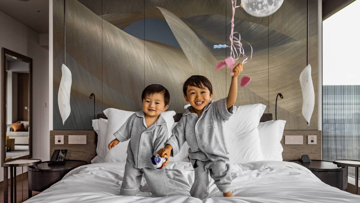 Two young children in grey pyjamas hold hands while standing on a bed in a bright room; one smiles and holds a clear balloon.